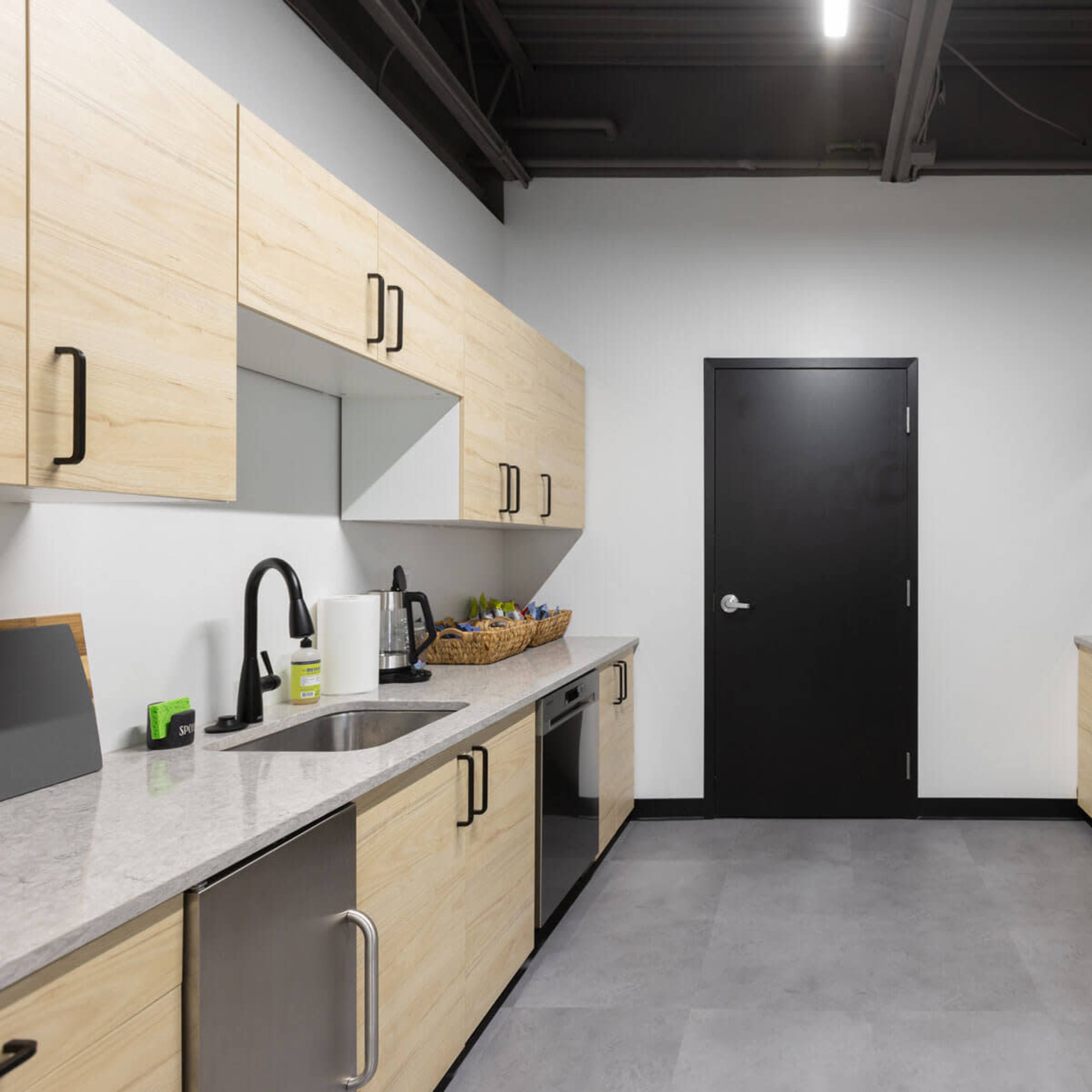 A modern kitchen with light wooden cabinetry, a stainless steel refrigerator, and a black door leading to another room.