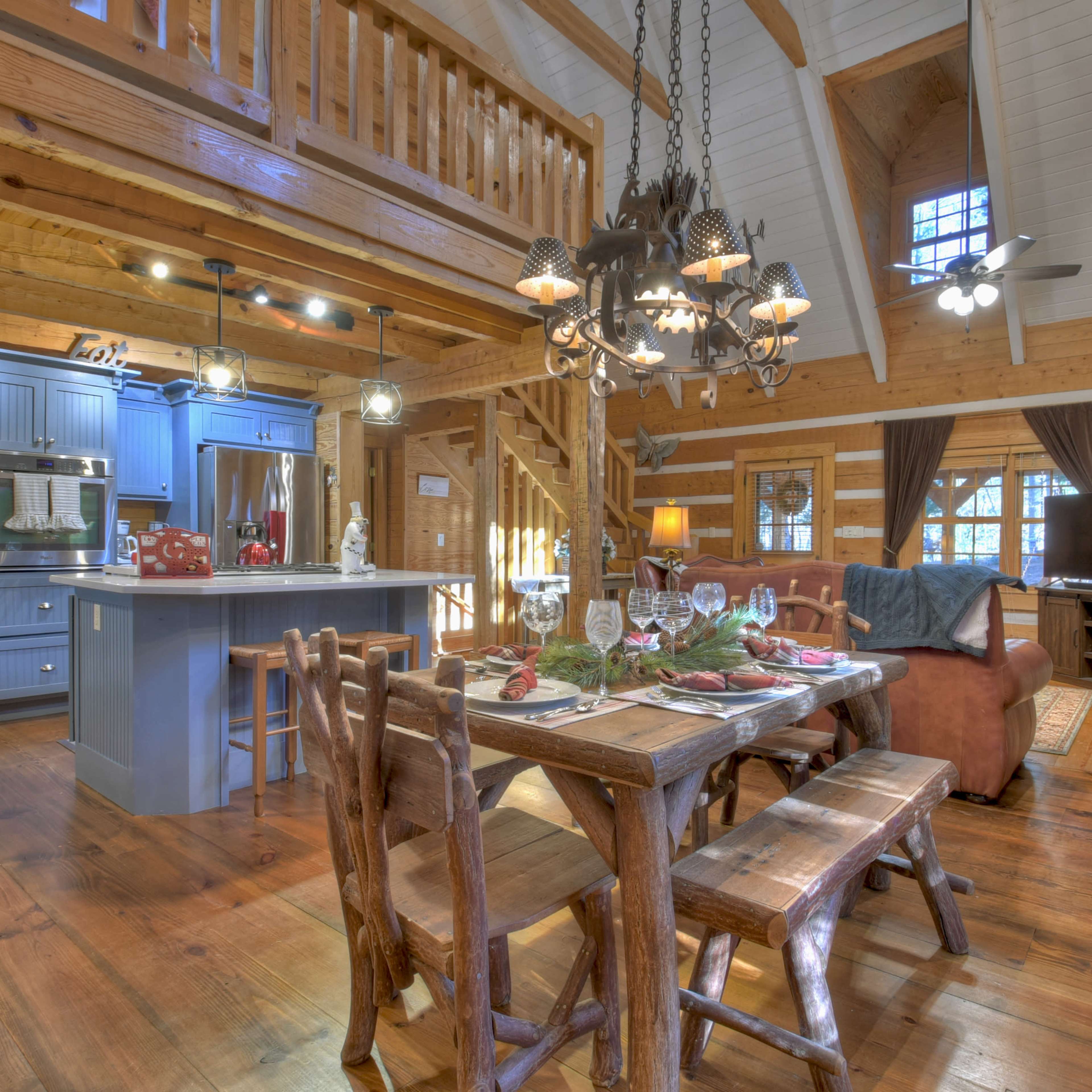 The interior of a rustic log cabin features a dining area with a wooden table, a kitchen with blue cabinetry, and a living area with a stone fireplace.