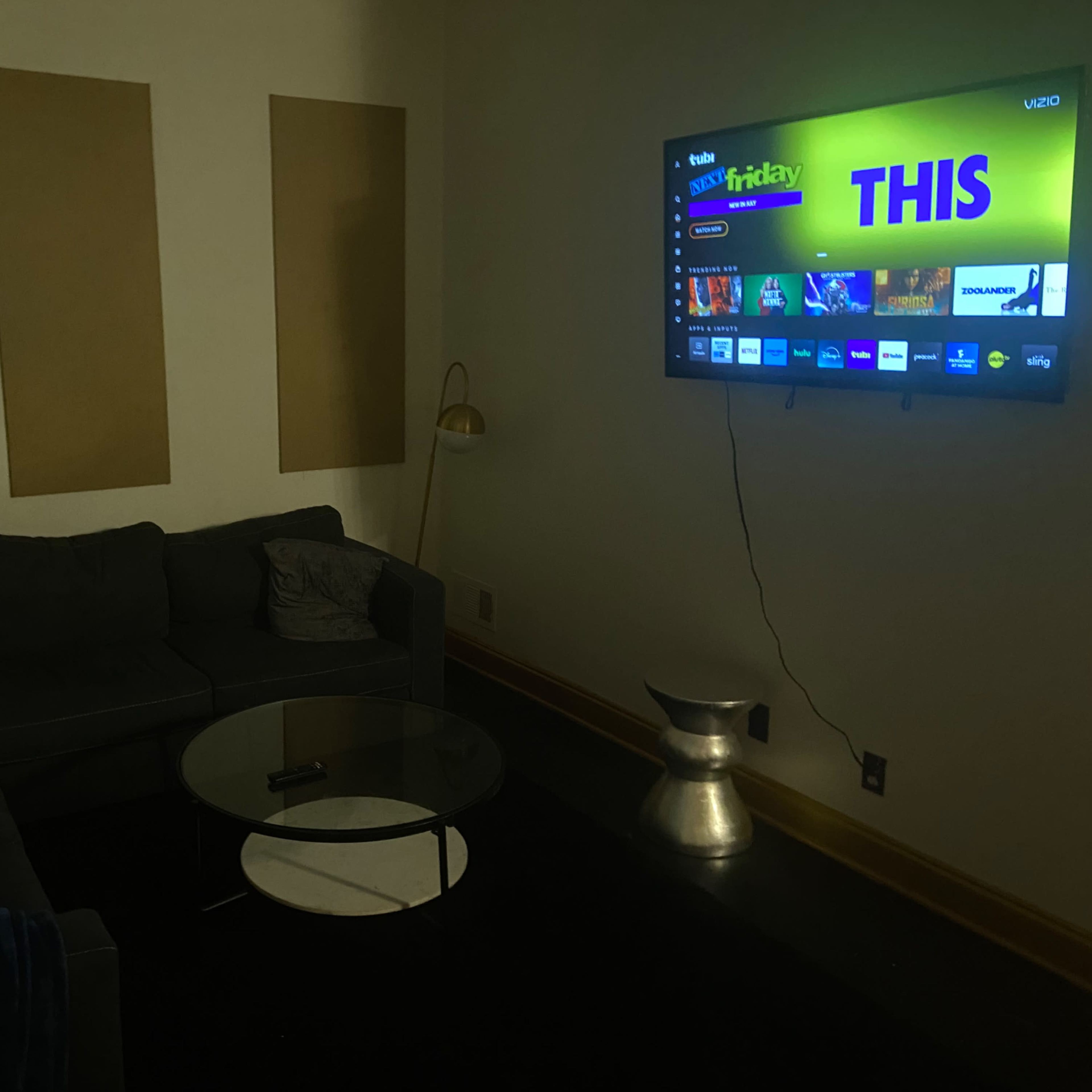 A dimly lit living room features a gray sectional sofa, a glass coffee table, and a wall-mounted TV displaying streaming content.