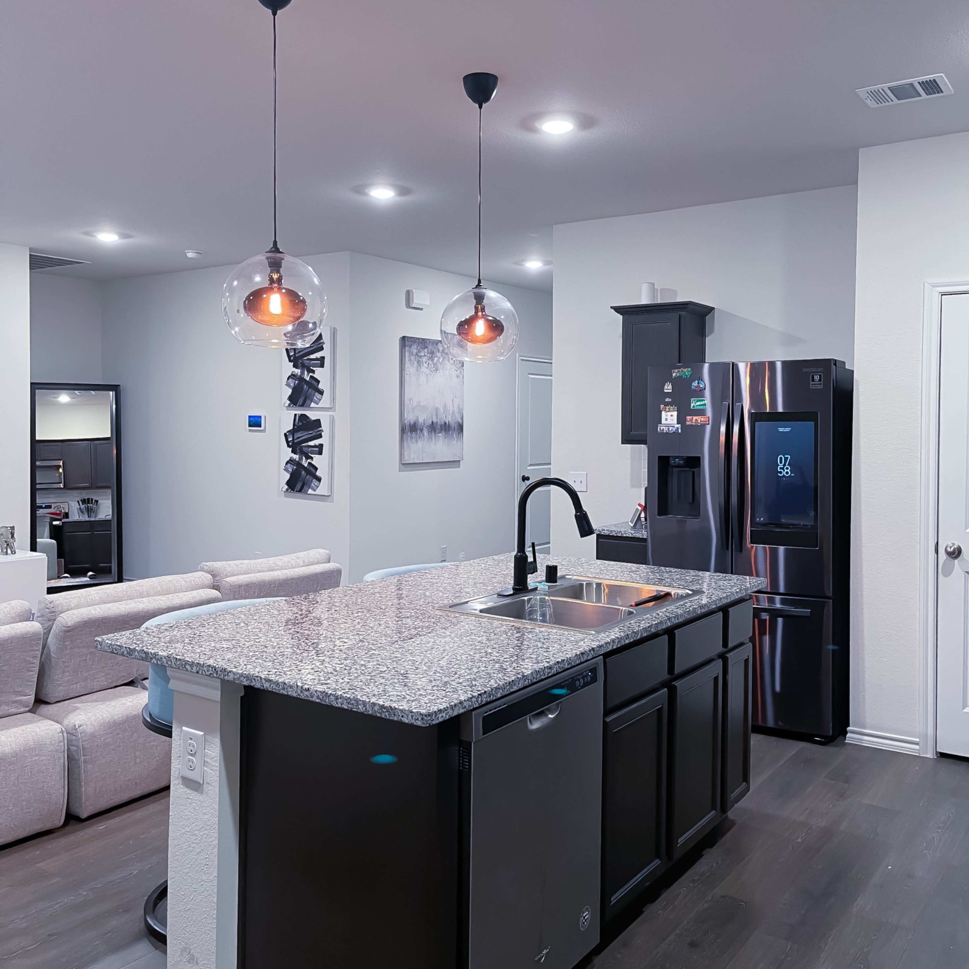 A modern kitchen features a large island with a sink and pendant lights, surrounded by a refrigerator and an open living area in the background.