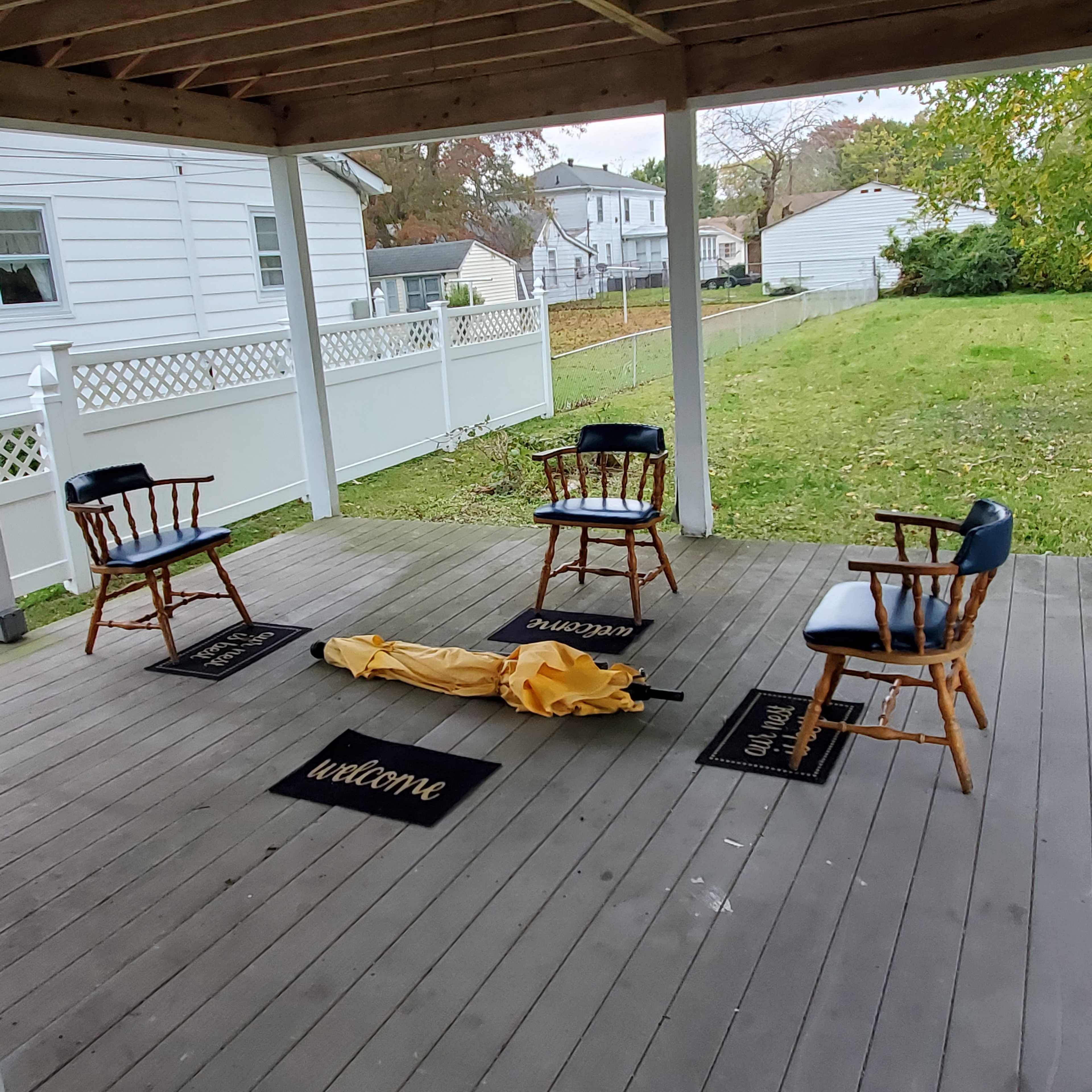 A yellow tarp lies on the ground surrounded by four wooden chairs on a porch with welcome mats.