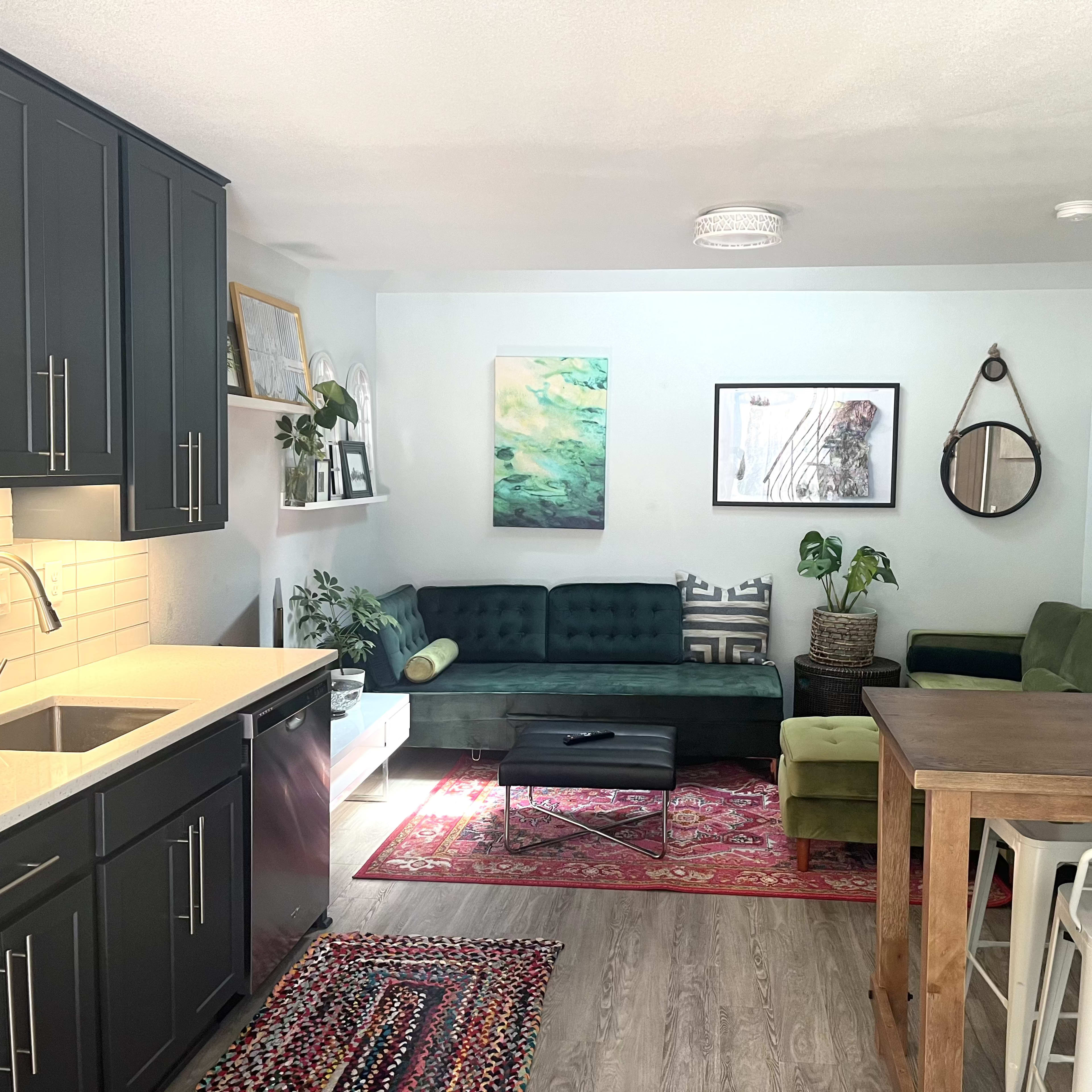 A modern living area features a green sectional sofa, a dining table, and a kitchen with dark cabinetry and a white backsplash.