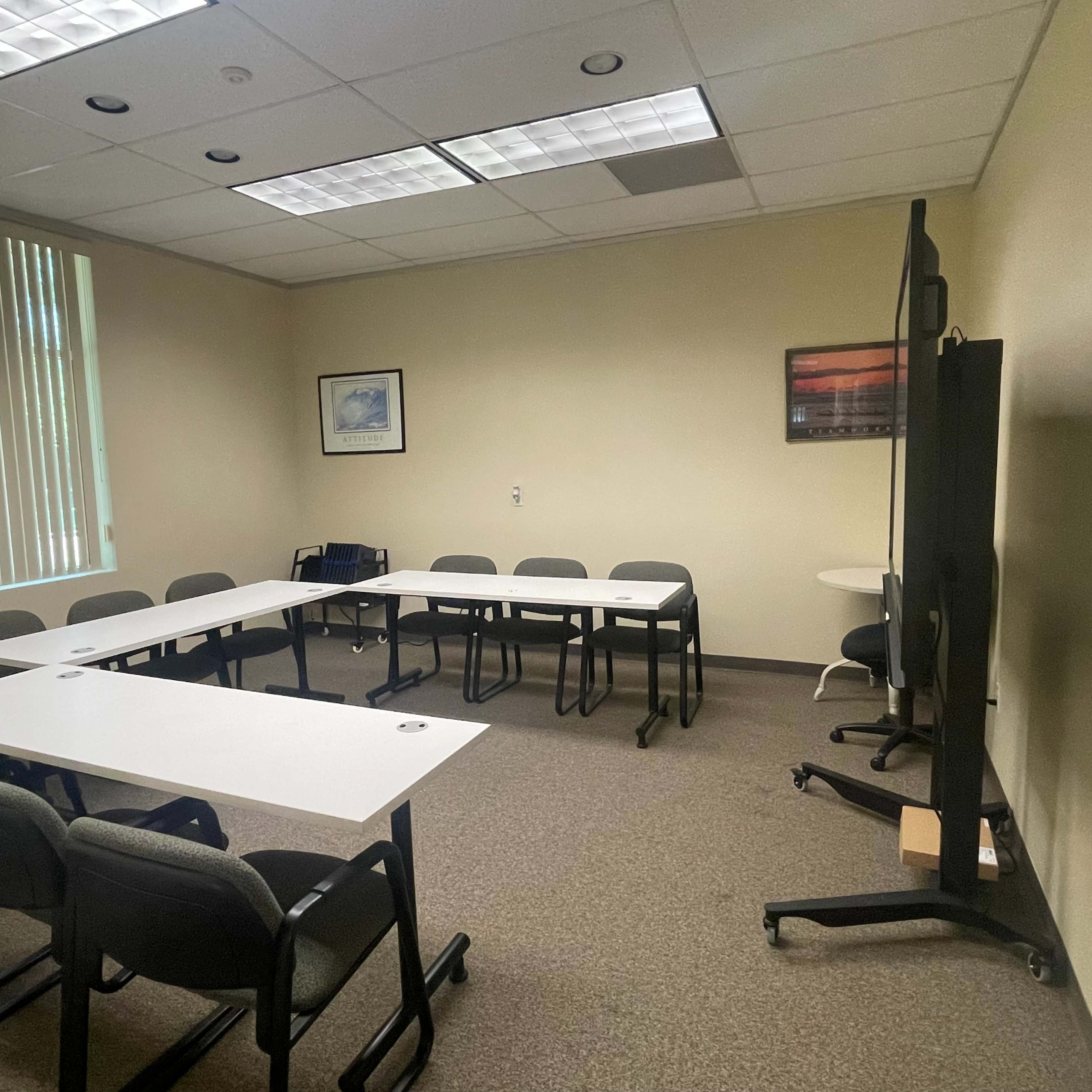 A conference room with several tables arranged in a U-shape and multiple chairs, along with a large screen on a stand.