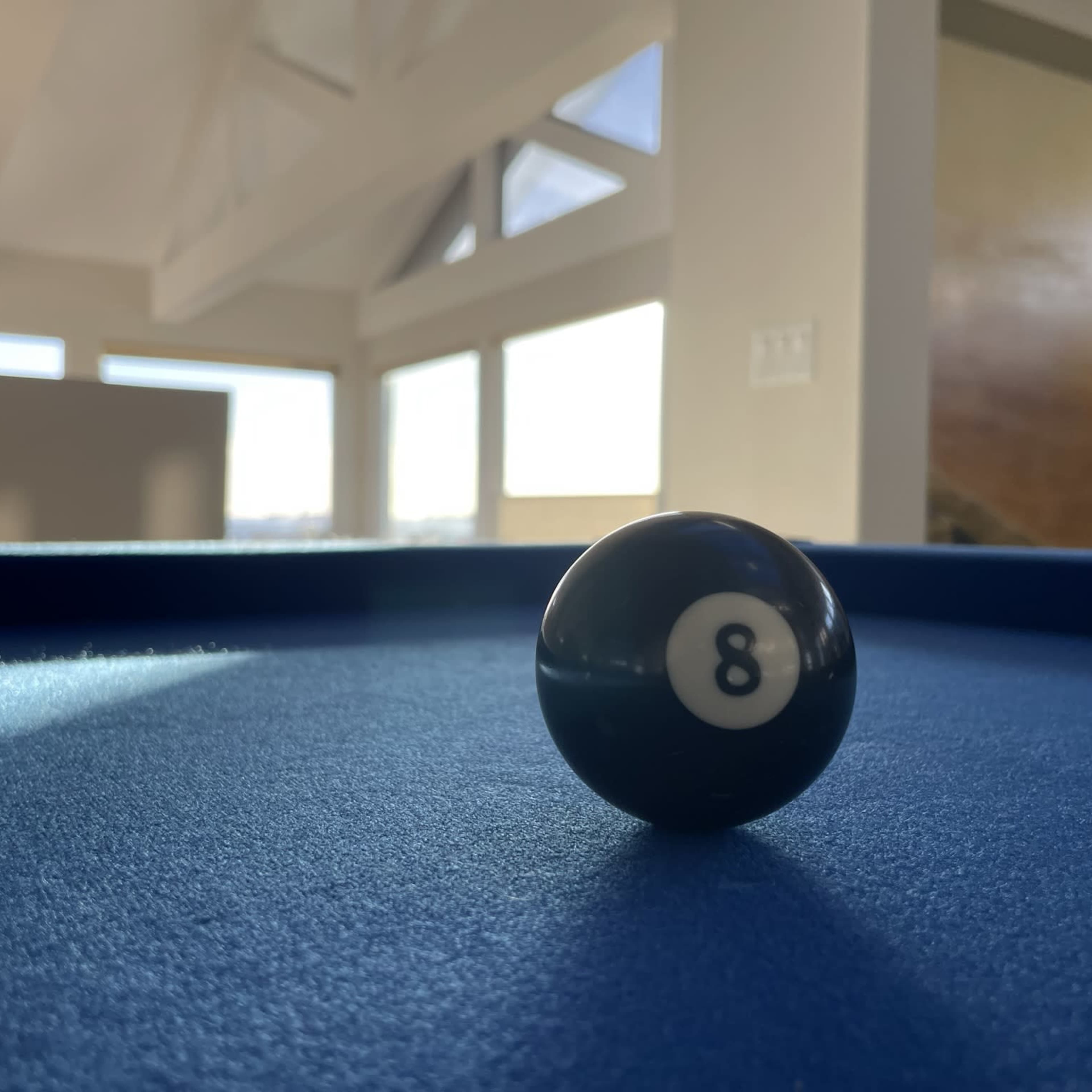 A black eight ball sits on a blue felt pool table with sunlight streaming through large windows in the background.