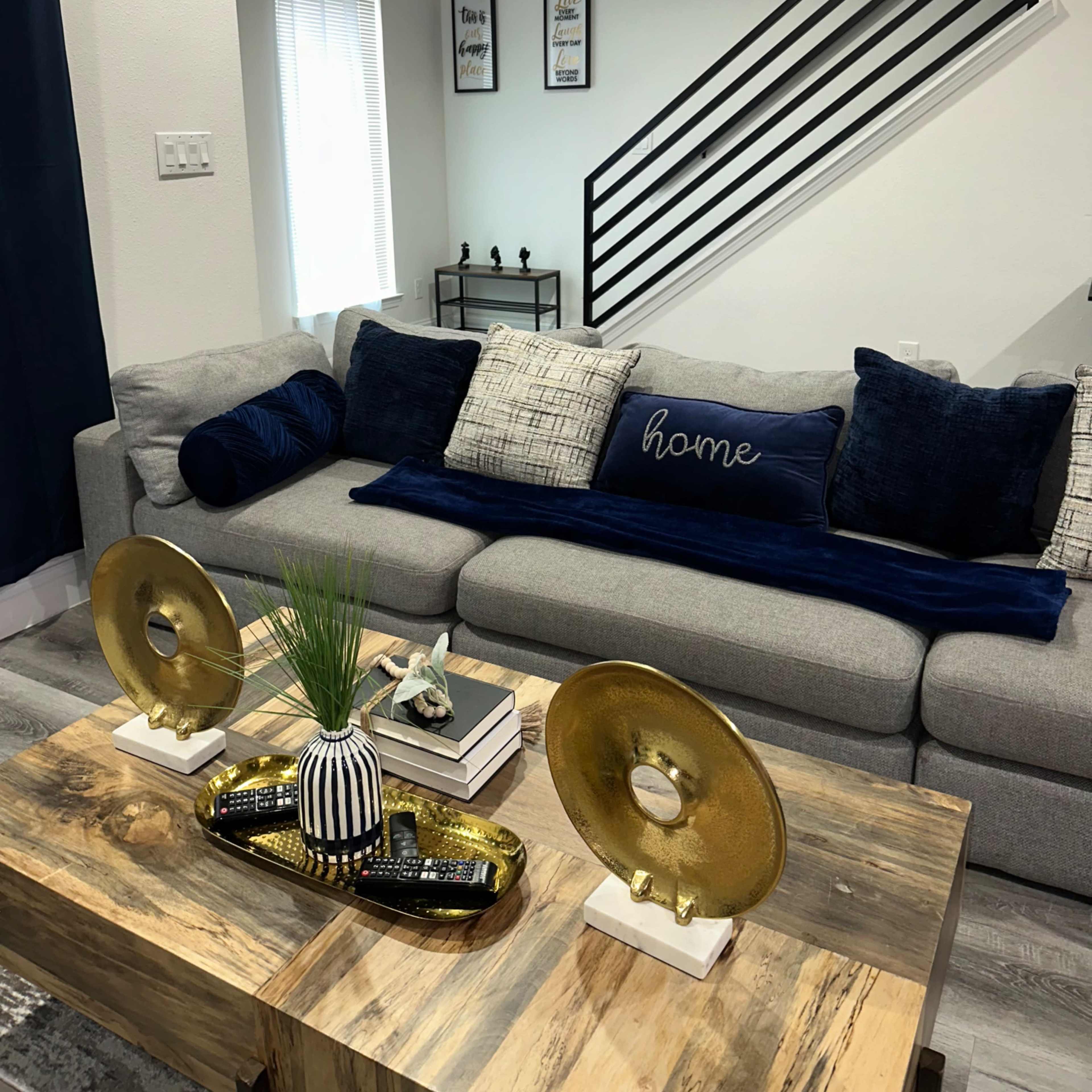 A modern living room featuring a gray sofa adorned with decorative pillows, a wooden coffee table with a decorative tray, and a staircase in the background.