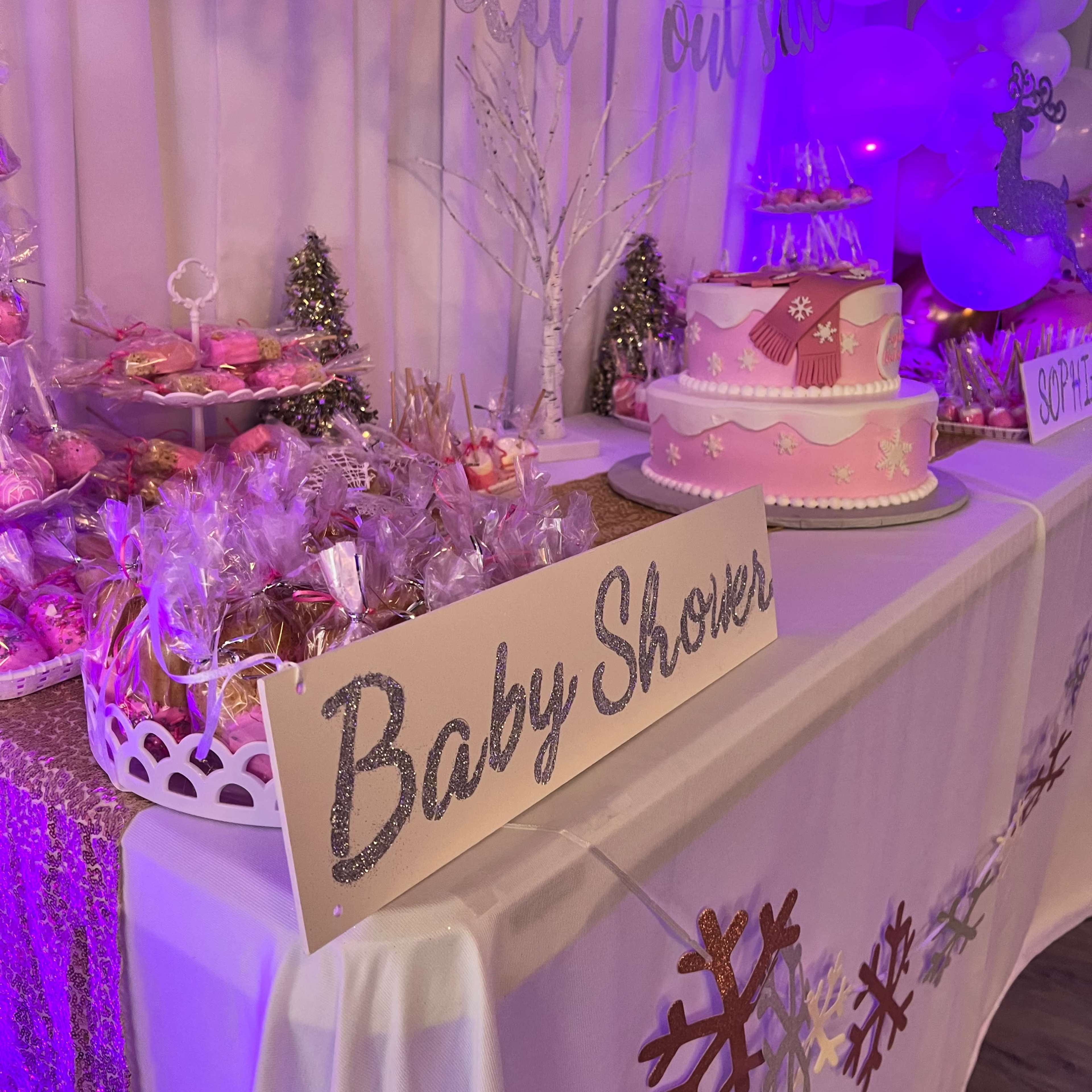 A pink and white cake is displayed on a table decorated for a baby shower, surrounded by treats and festive decorations.