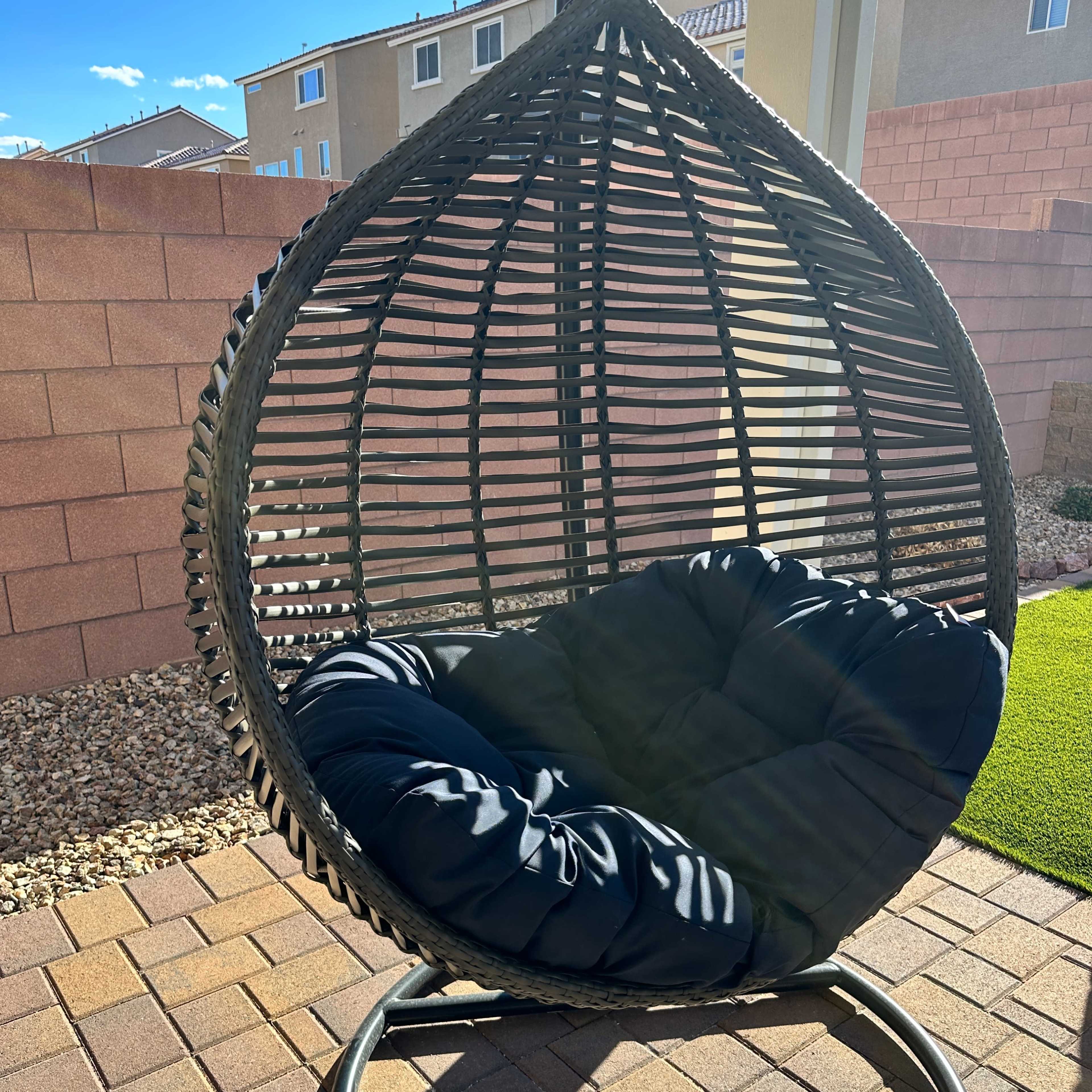 A large, teardrop-shaped hanging chair with a black cushion is positioned on a patio with stone pavers, surrounded by a low wall and grass.