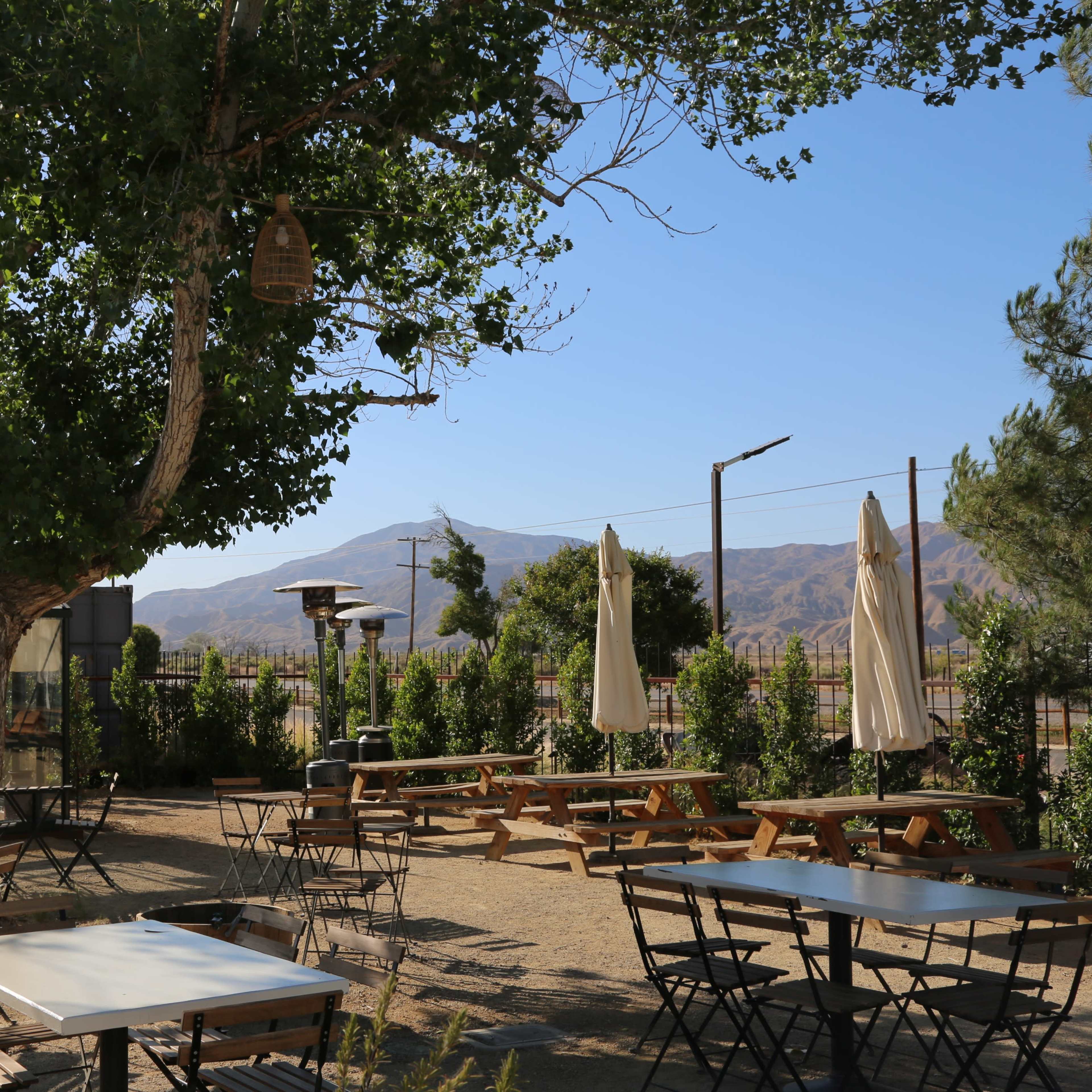 A patio with wooden tables and chairs is set up under a large tree, with mountains visible in the background.