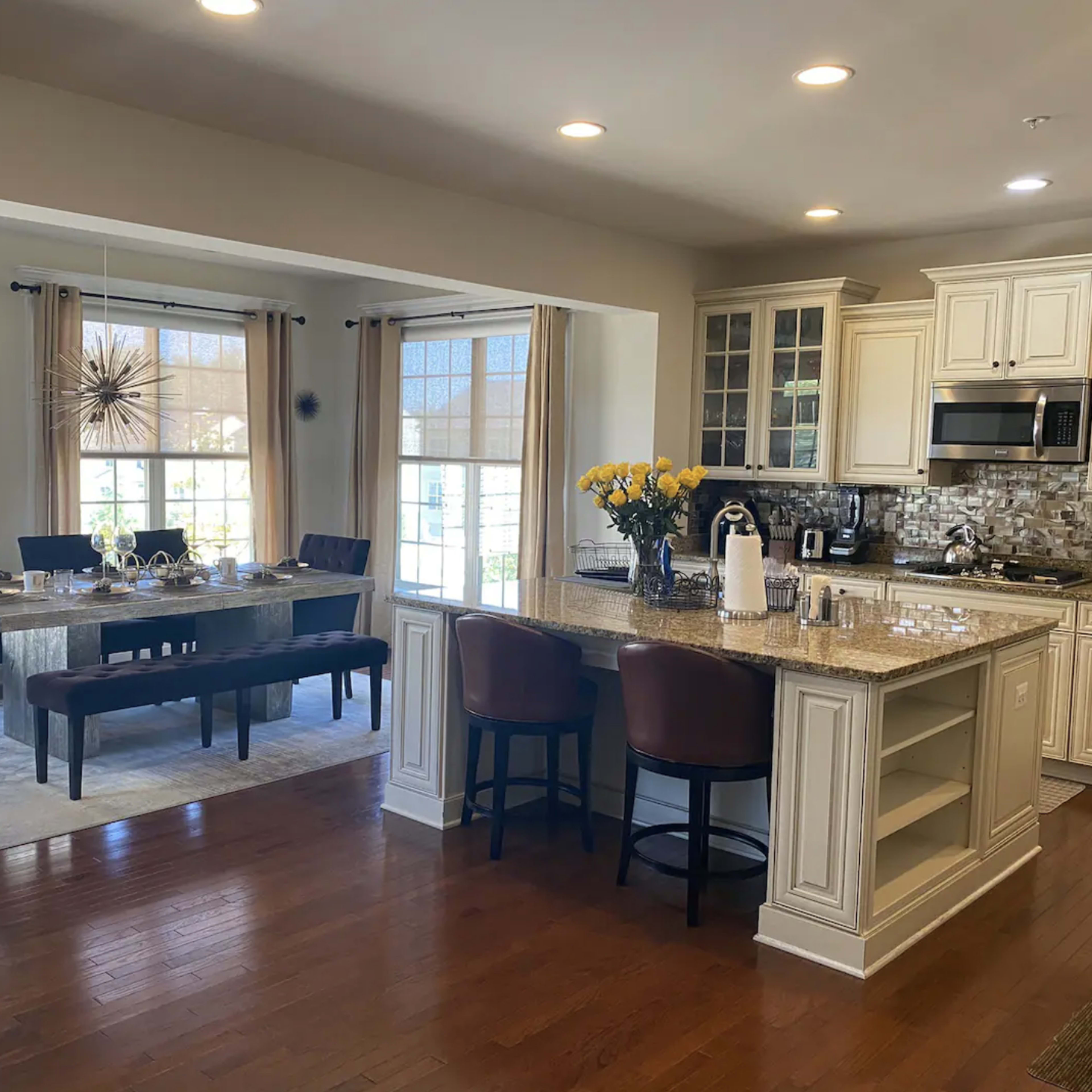 A modern kitchen and dining area feature a large table set for a meal, with natural light illuminating the space through multiple windows.