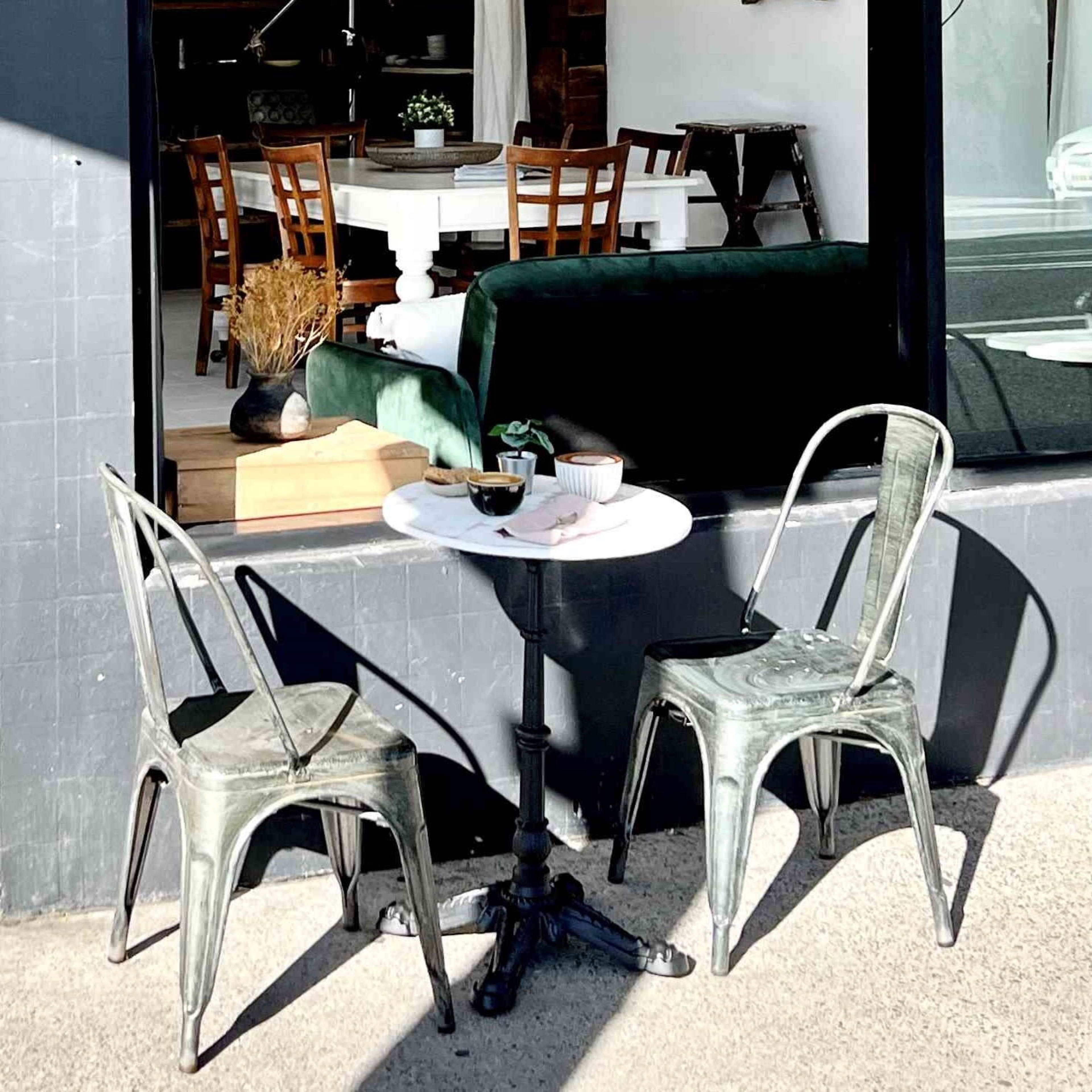A set of metal chairs and small round tables sit outside a café next to a large window reflecting the interior space and street.