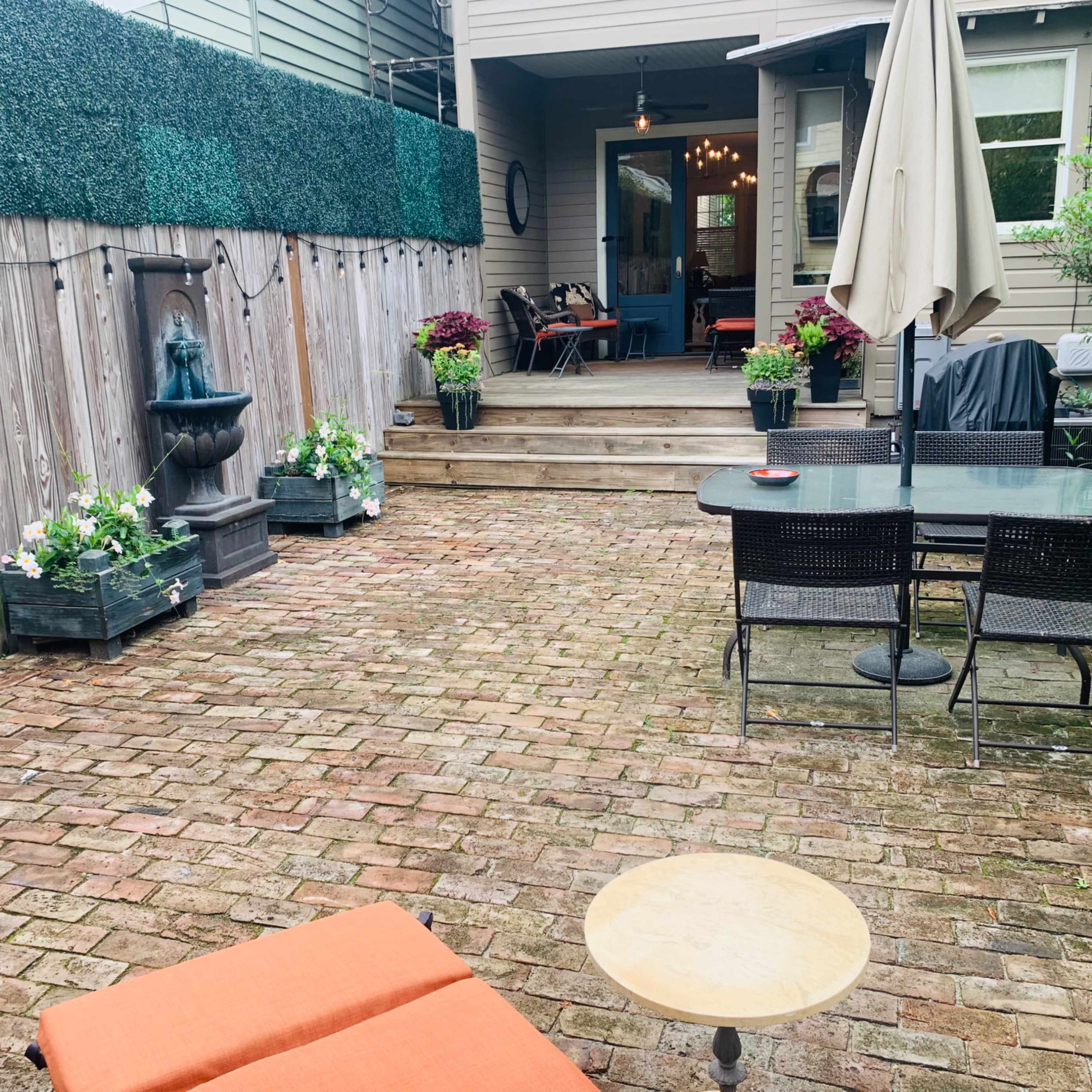 A brick patio featuring a small table and chairs, a fountain, and planters filled with flowers, leading to a door that opens into a home.