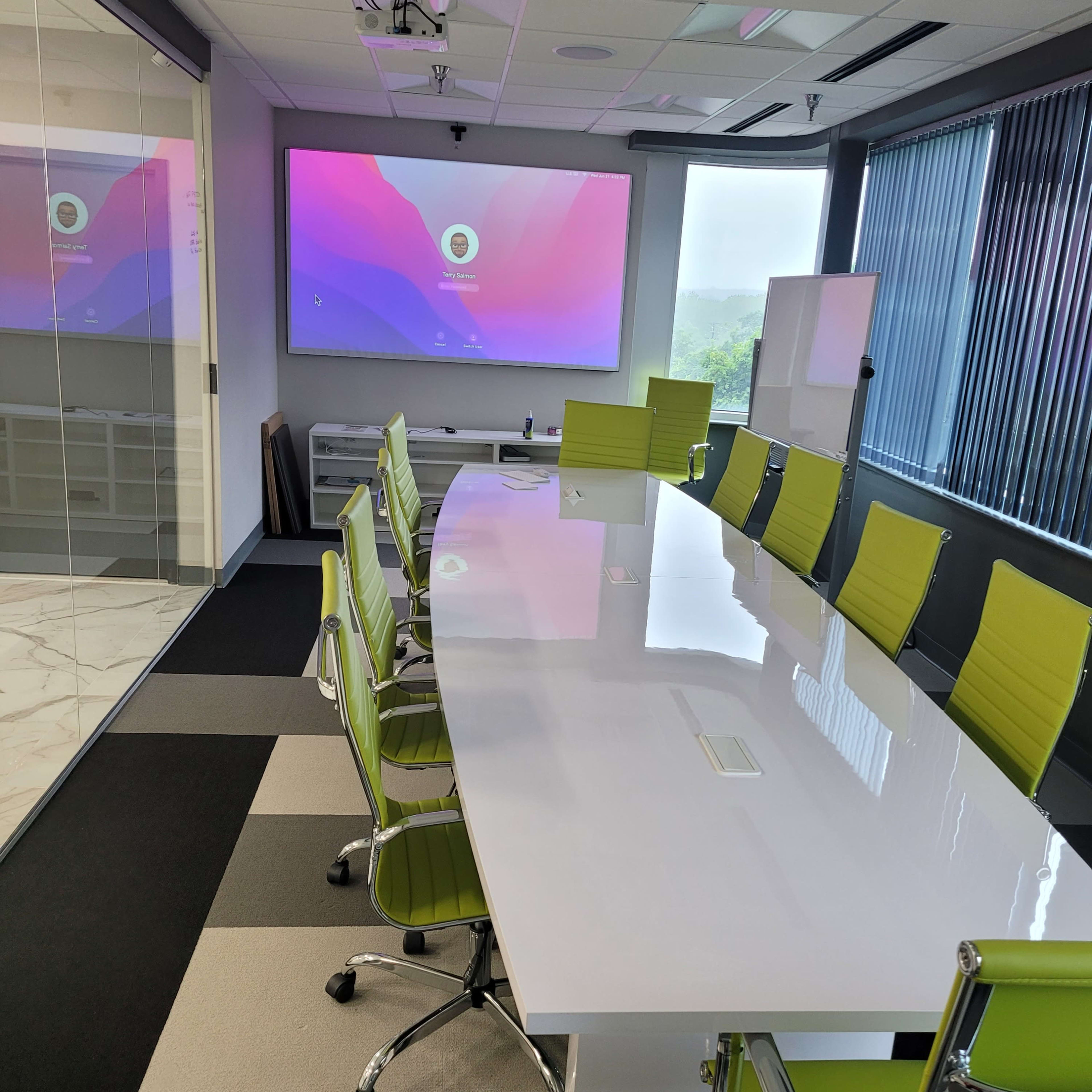 A modern conference room features a long white table surrounded by green chairs, with a large screen displaying a colorful presentation in the background.