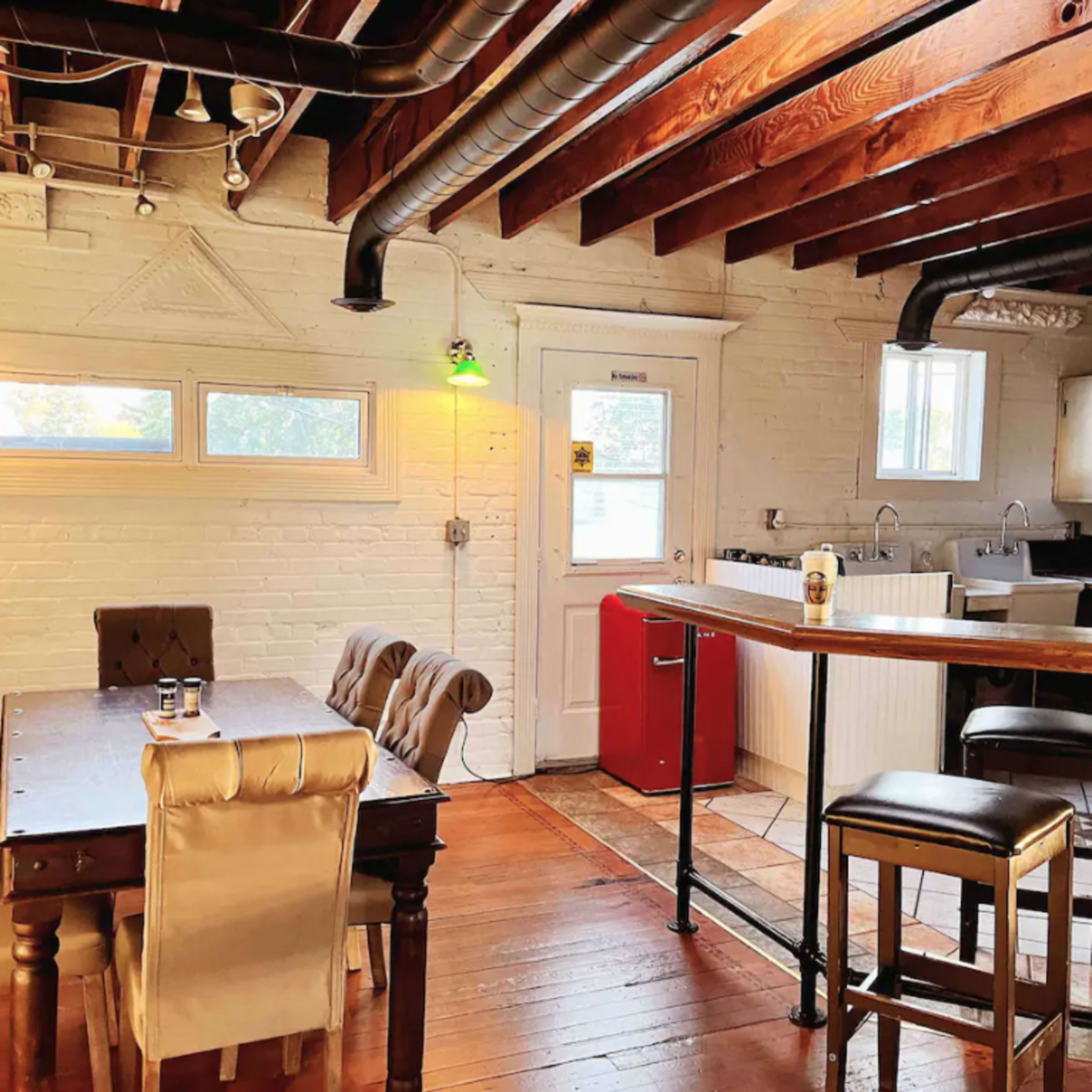 The scene depicts a rustic kitchen area featuring a wooden dining table with upholstered chairs, a bar-height countertop, and exposed beams on the ceiling.