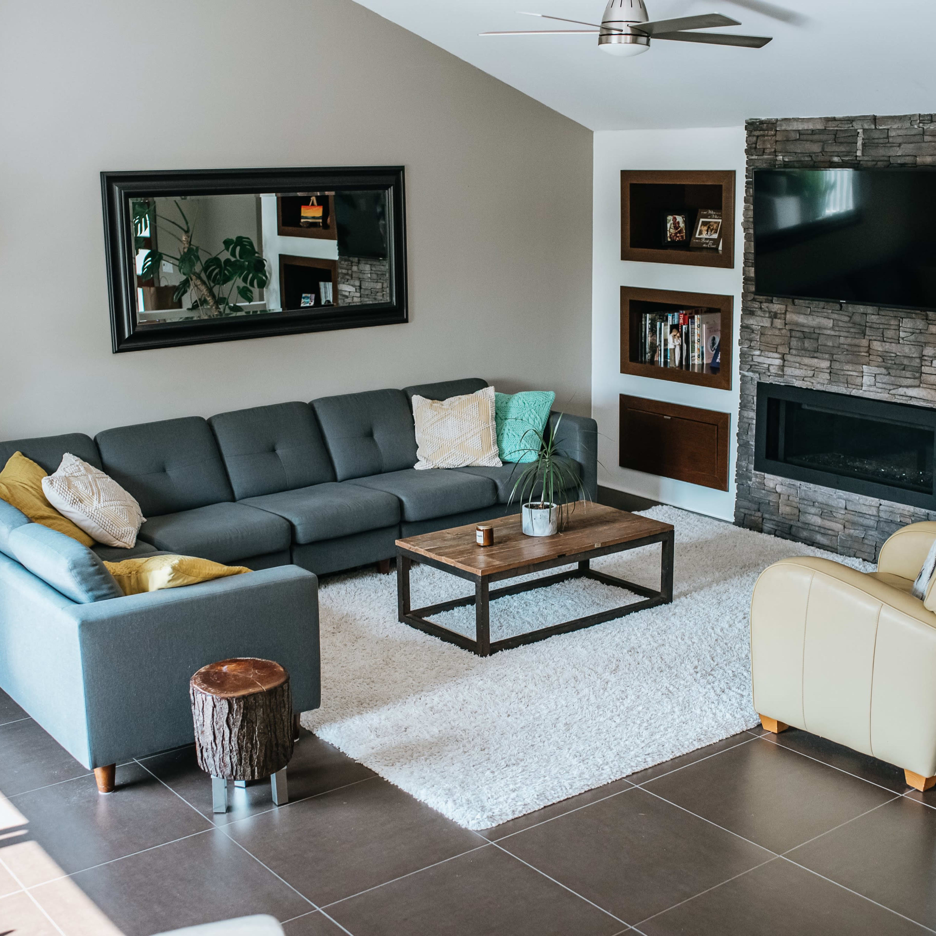 A modern living room features a gray sectional sofa, a wooden coffee table, a stone fireplace, and shelves with books and decorative items.