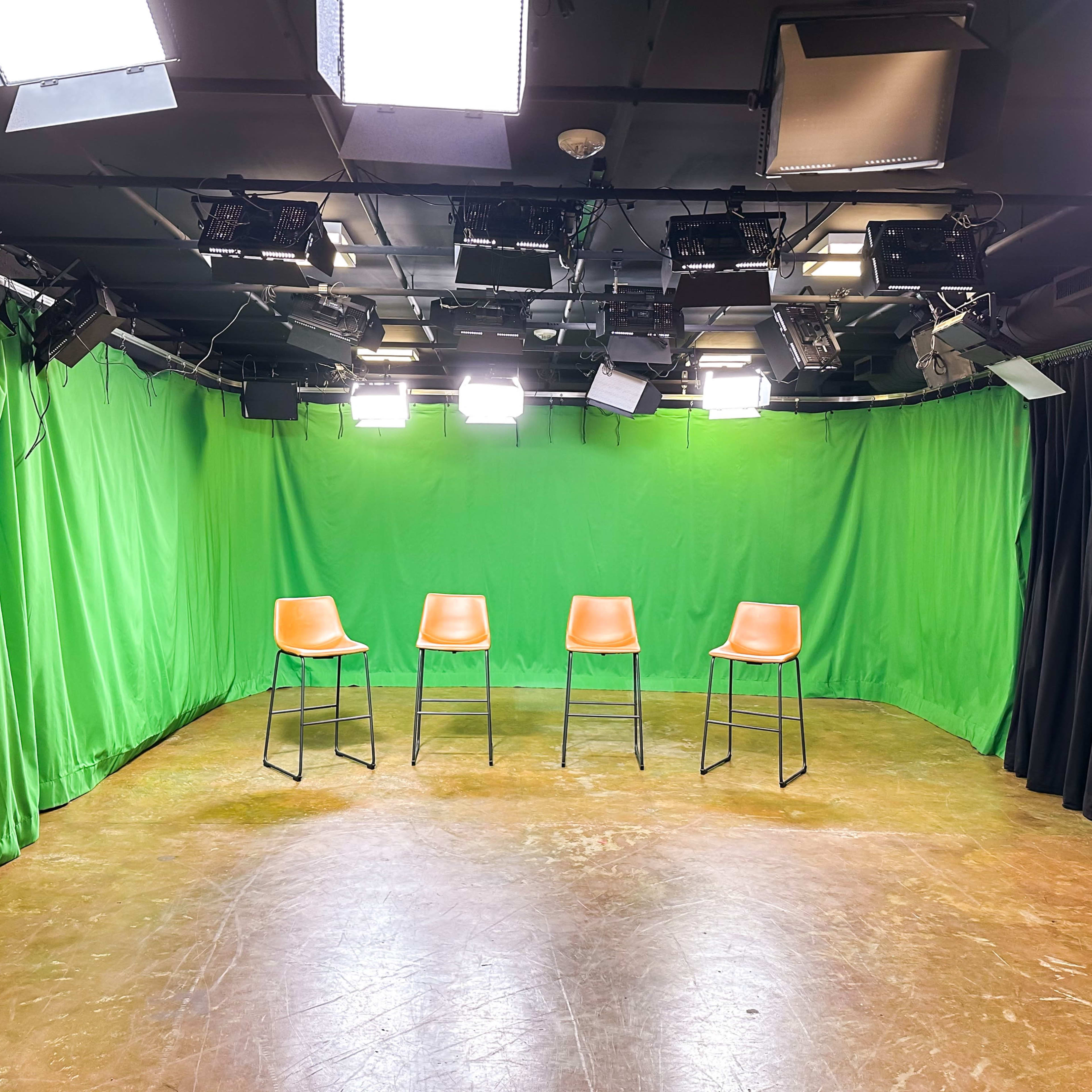 A television studio setup with four empty orange bar stools on a bare floor surrounded by green and black curtains and bright overhead lights.