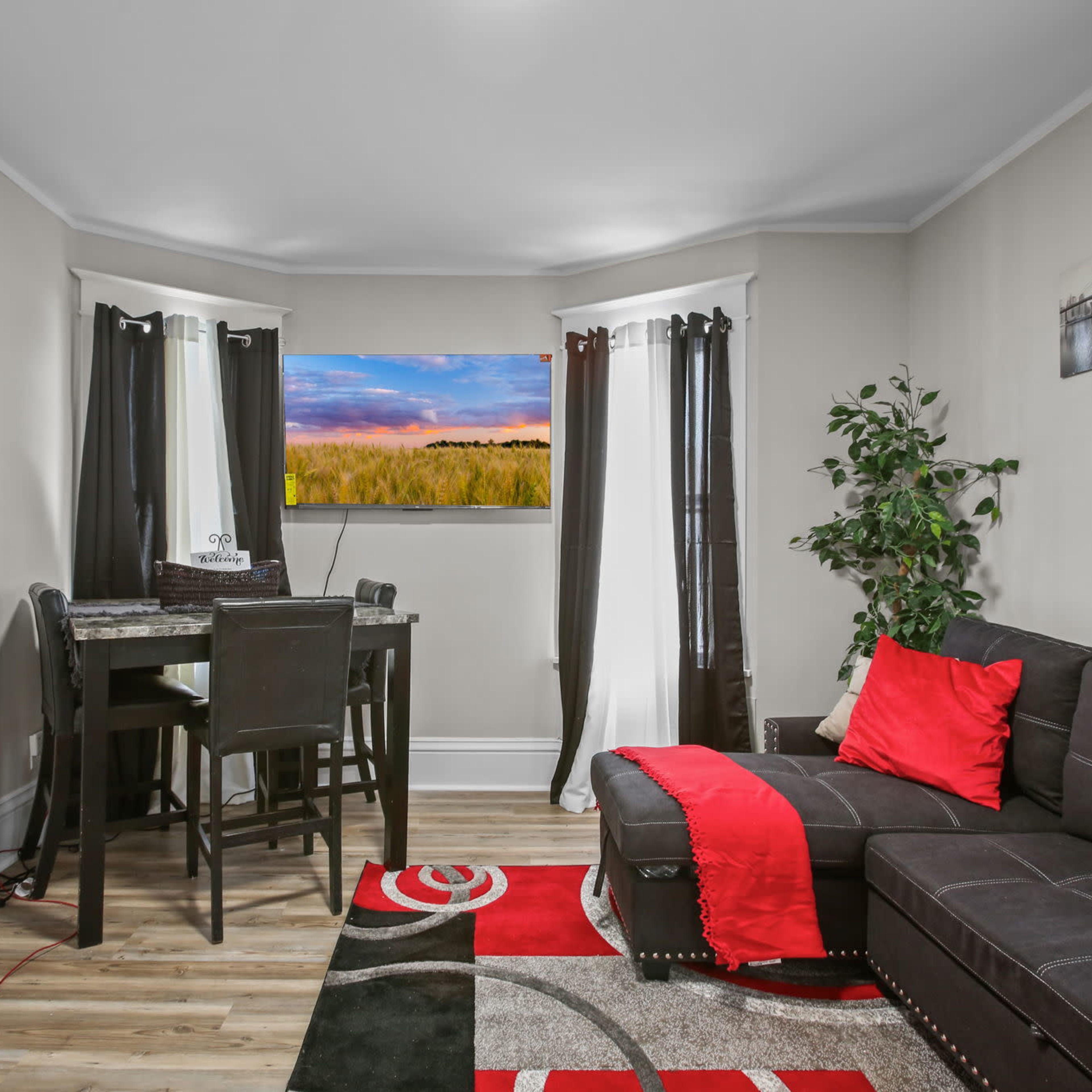 A small, modern living space features a black sectional sofa, a dining table with chairs, and a colorful area rug.