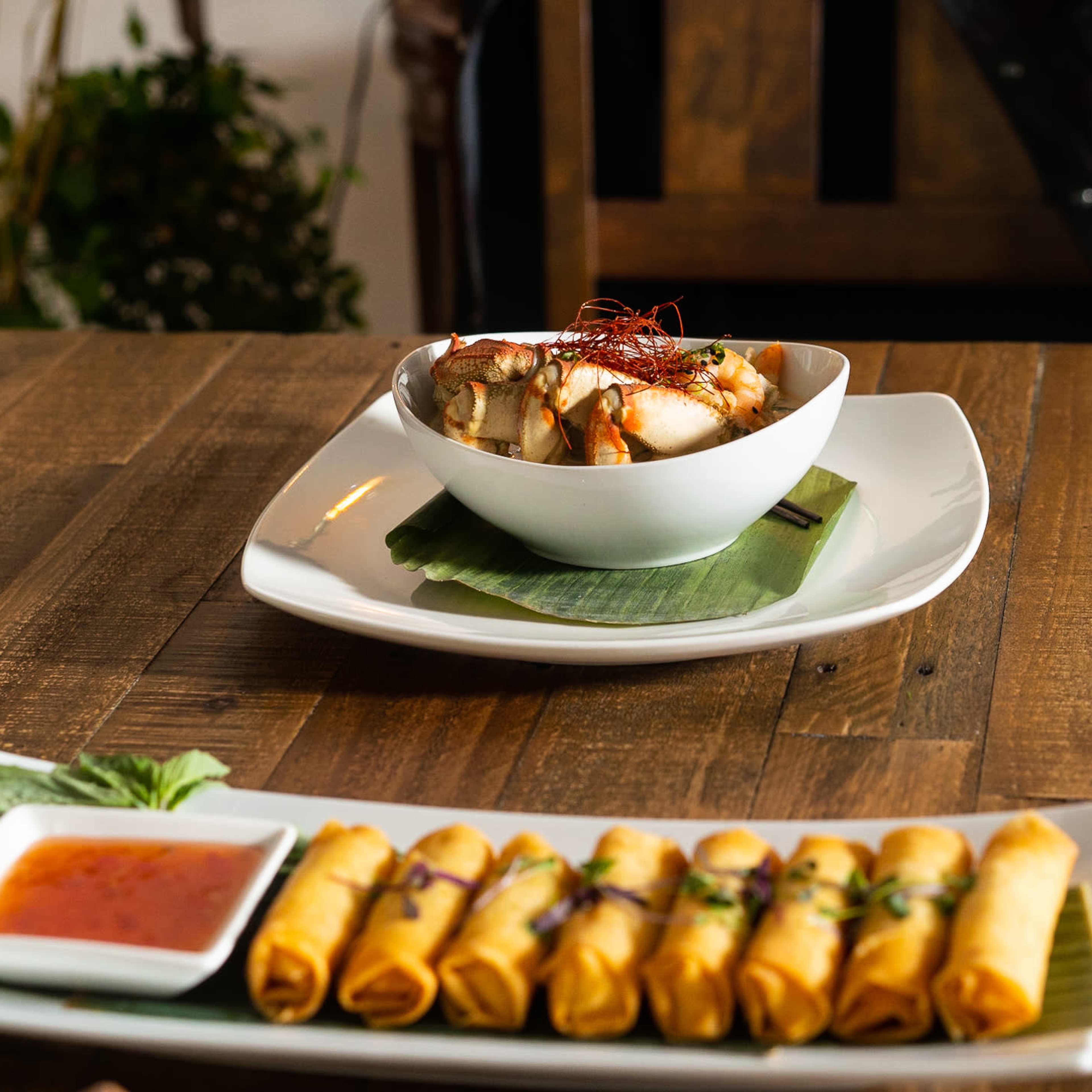 A plate of spring rolls with dipping sauce sits in front of a bowl of shrimp on a wooden table.