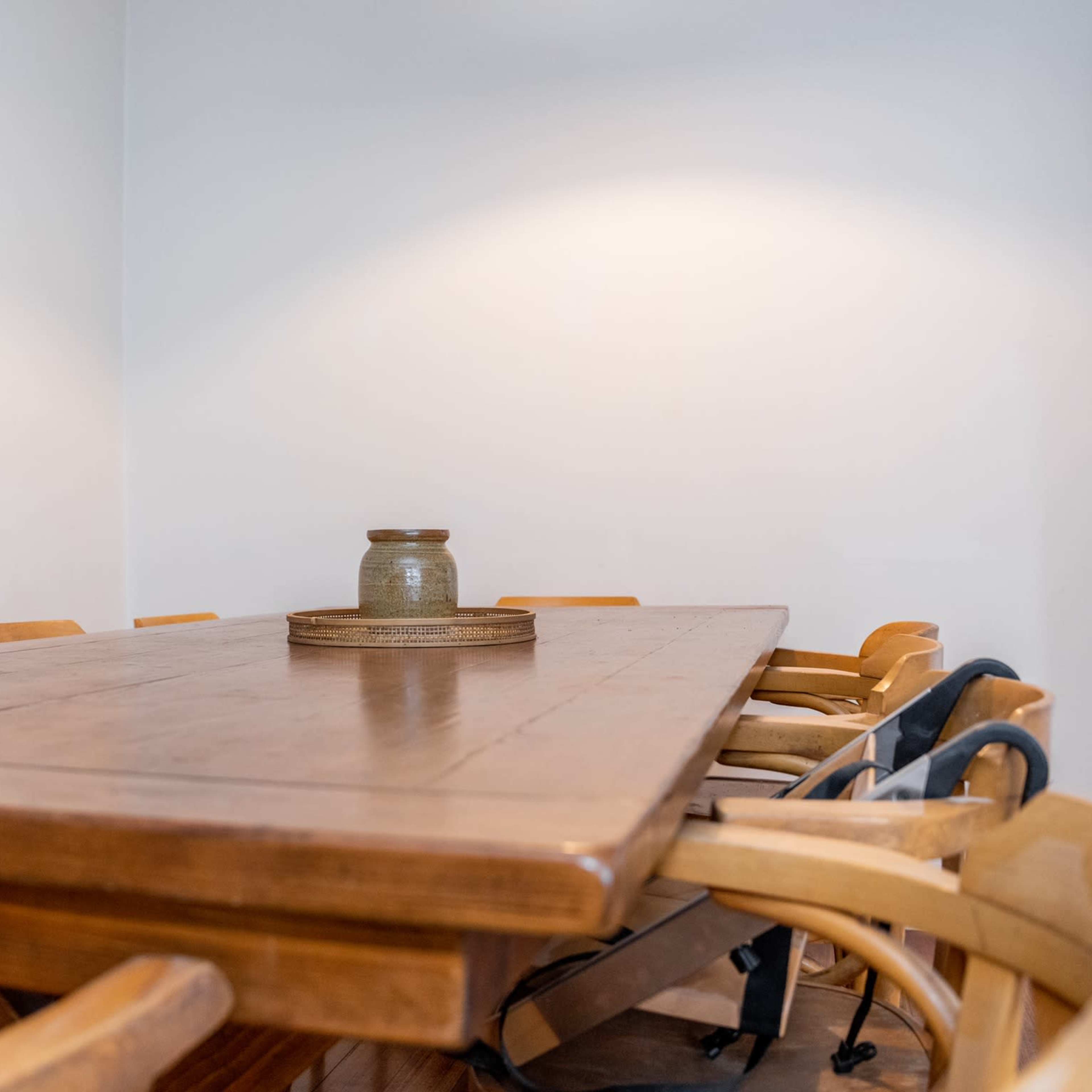 A wooden dining table with a ceramic vase in the center is surrounded by several wooden chairs in a sparsely decorated room.