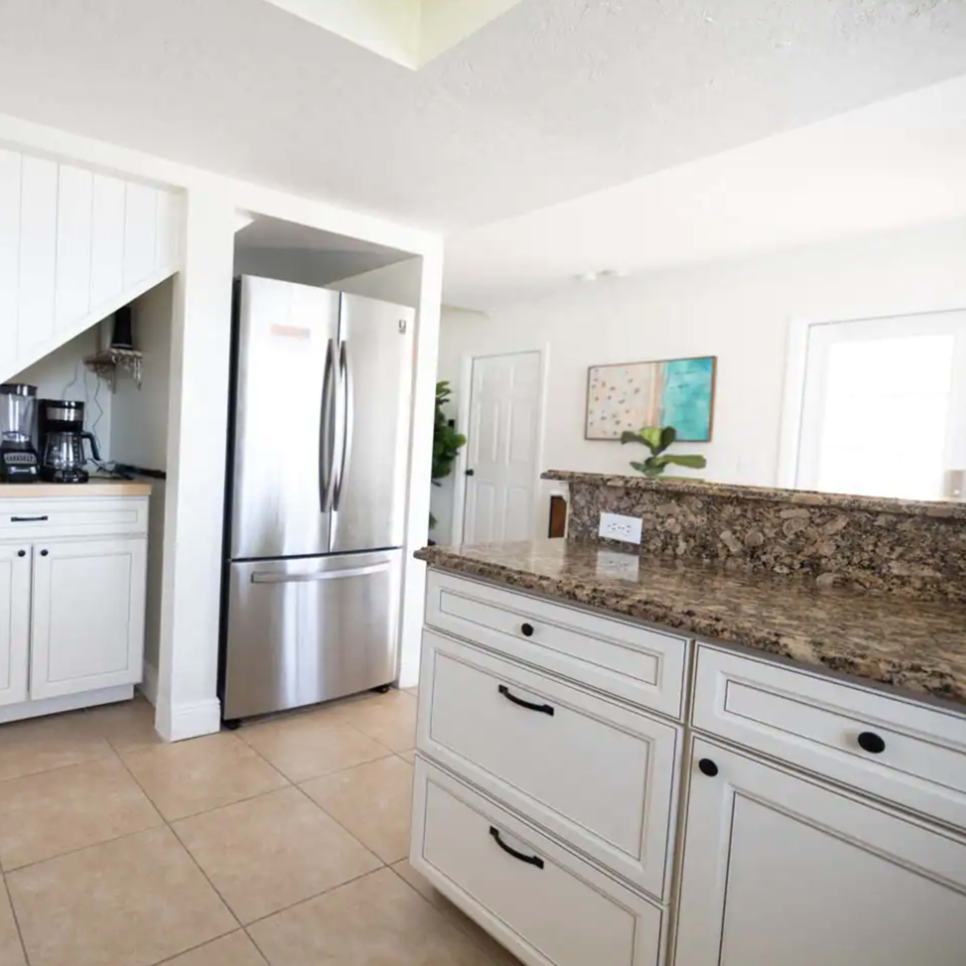 The kitchen features white cabinetry, granite countertops, and stainless steel appliances, with a coffee maker positioned in an alcove under the stairs.