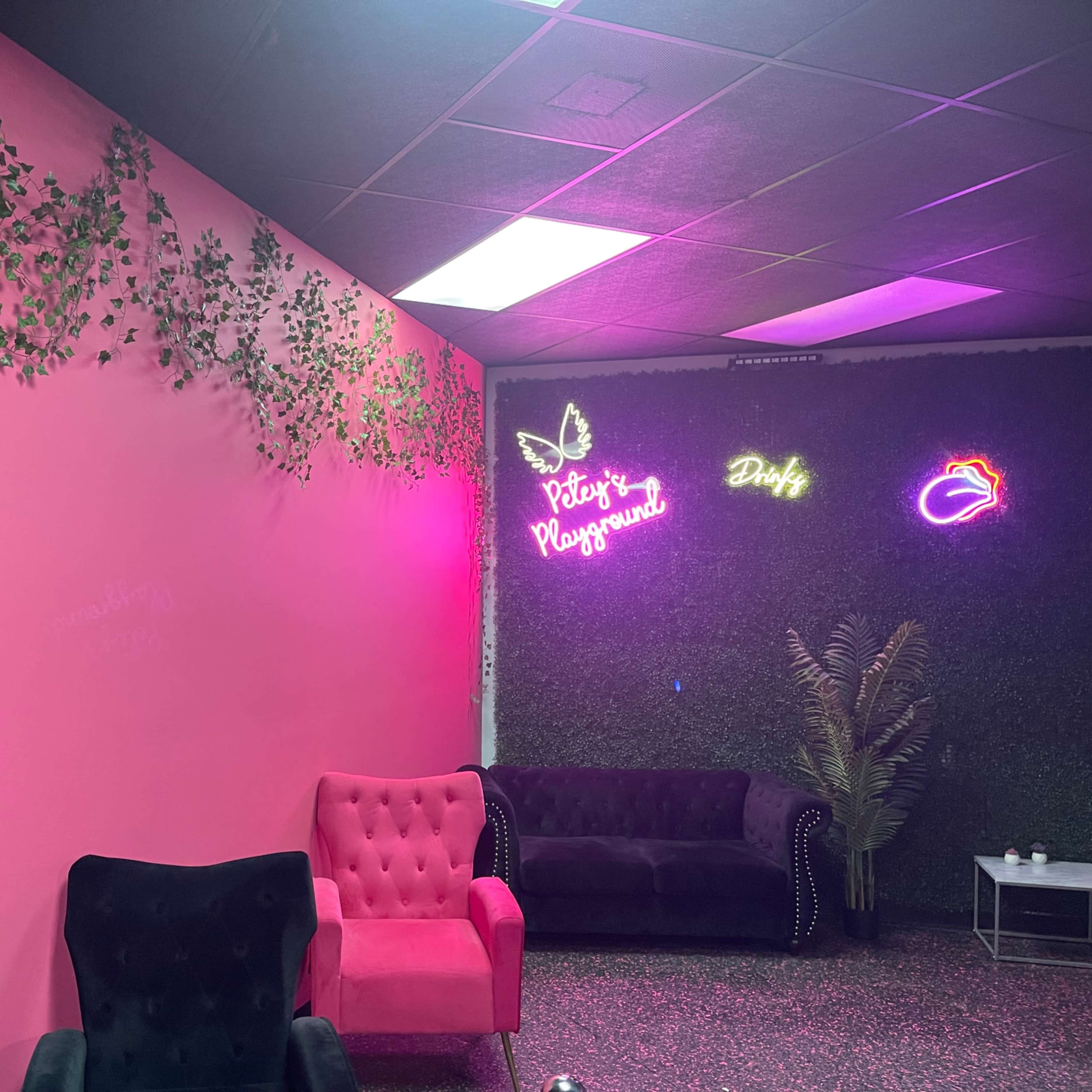 The scene features a brightly colored pink wall adorned with green vines, against which neon signs and plush seating are arranged in a stylish interior.