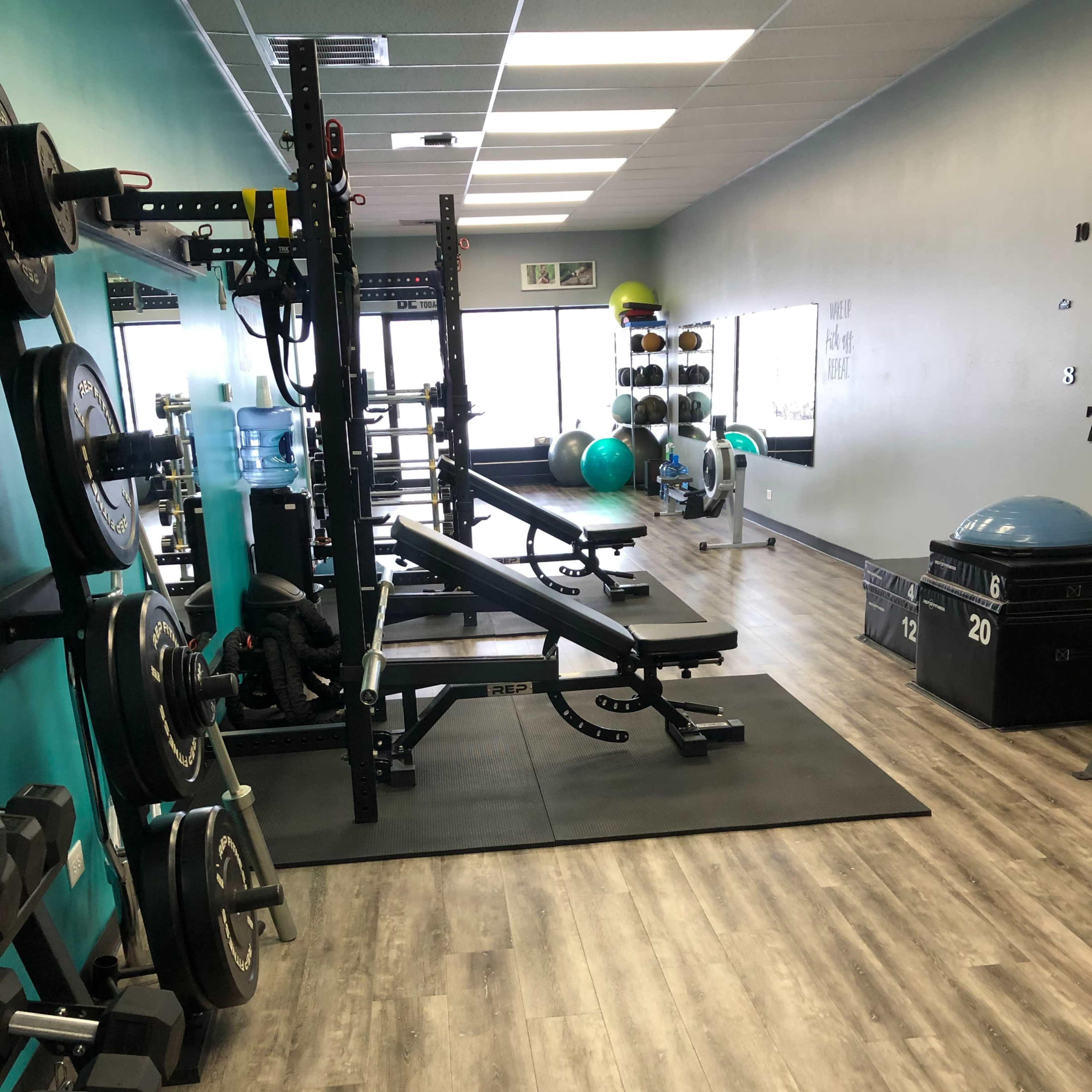 A fitness gym interior with various workout equipment, including weights, benches, and exercise balls, along a well-lit space.