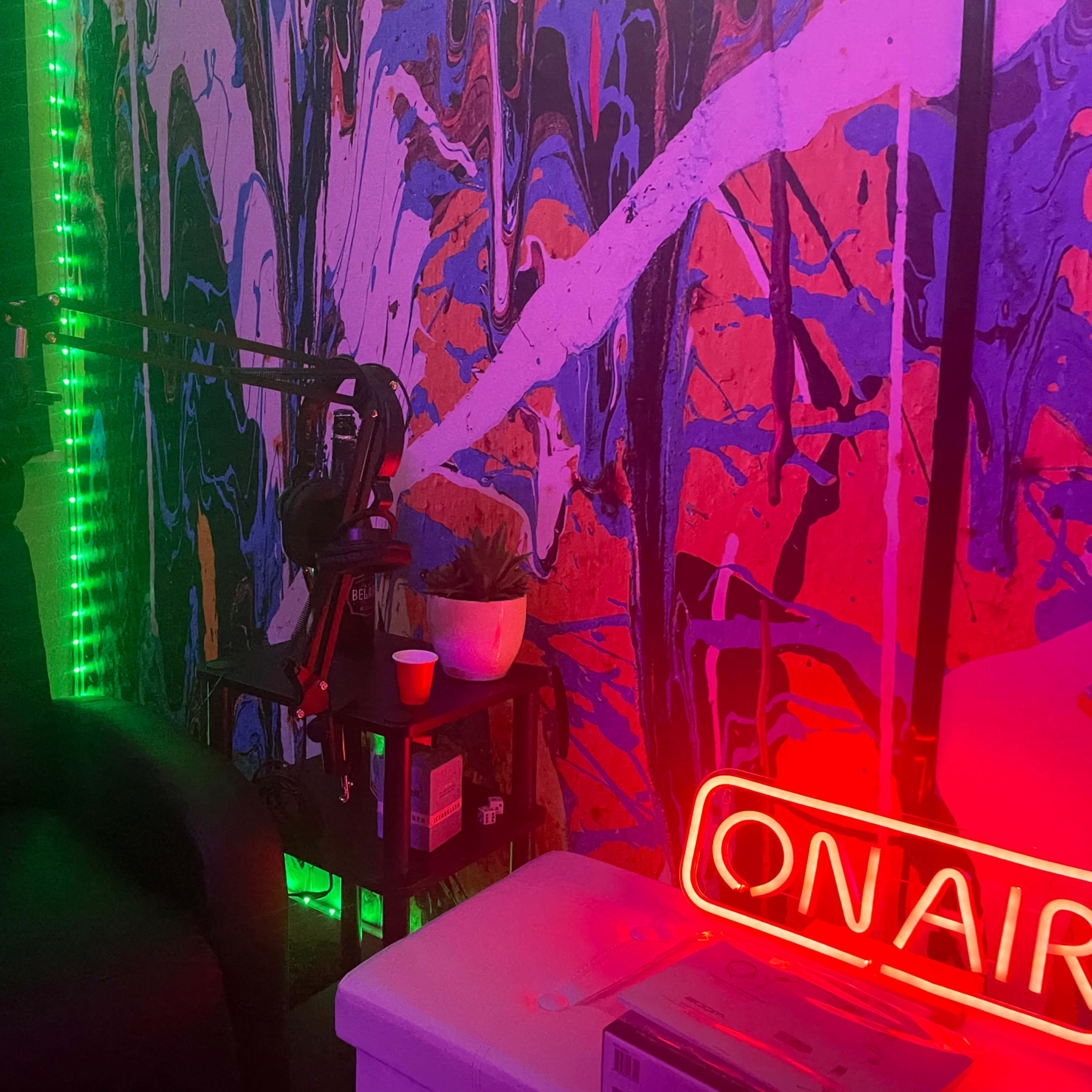 A corner of a recording studio with a chair, a microphone on an adjustable arm, a small table with a plant and cup, and vibrant neon lights along the walls.