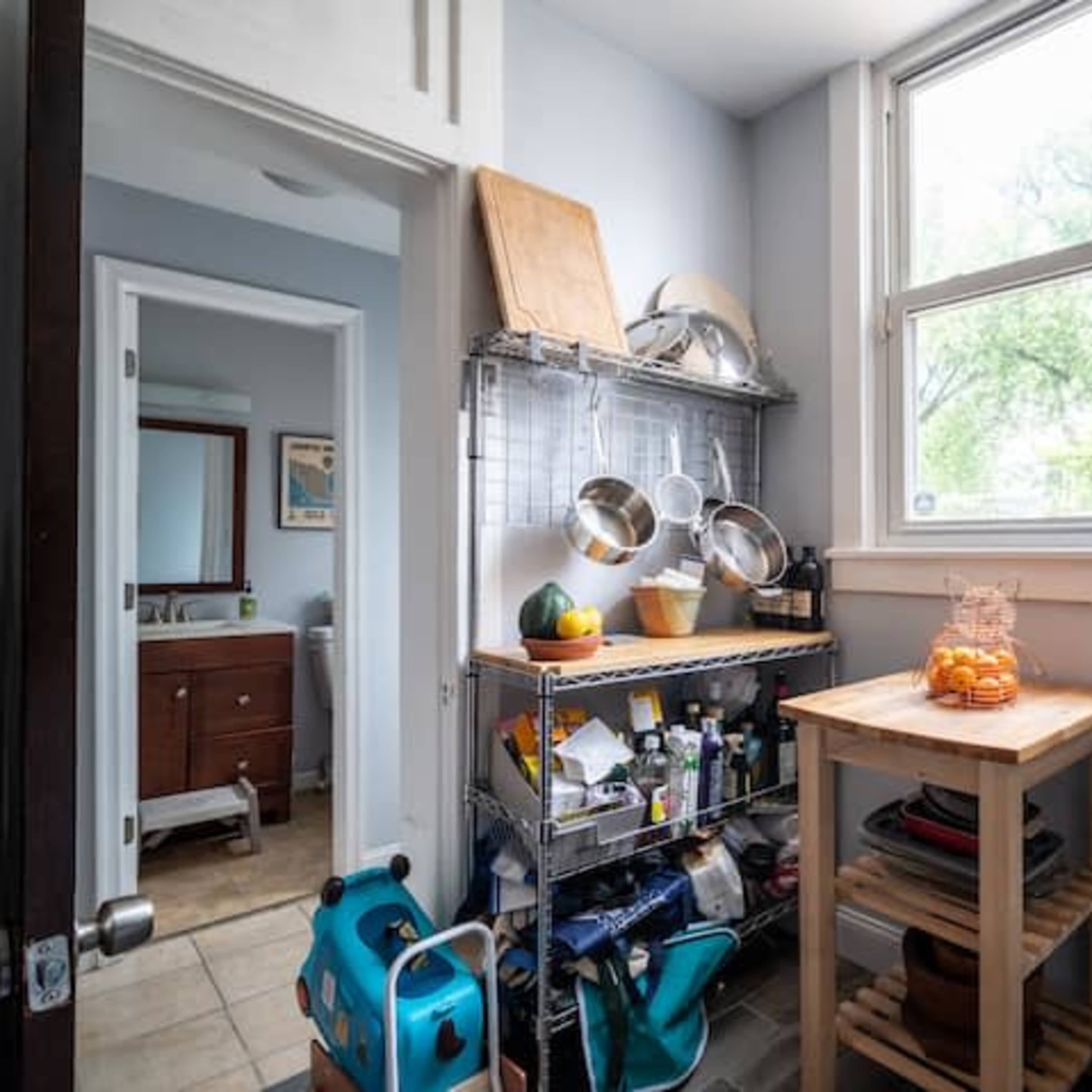 A small kitchen space featuring a metal shelving unit with cooking utensils and a wooden table, leading to a bathroom area.