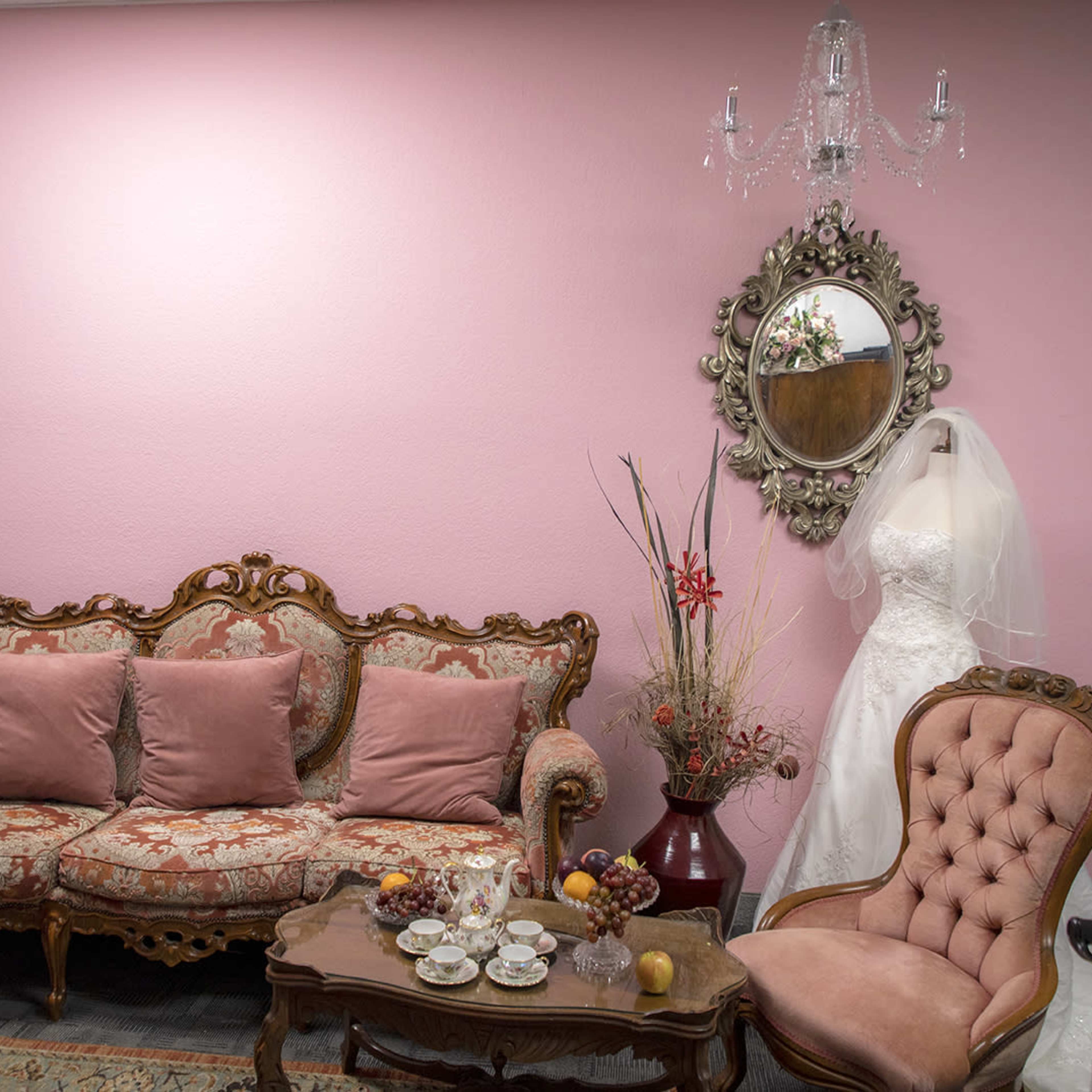 A room features a pink wall, vintage furniture with floral upholstery, and a wedding dress displayed on a hanger beside a mirror.