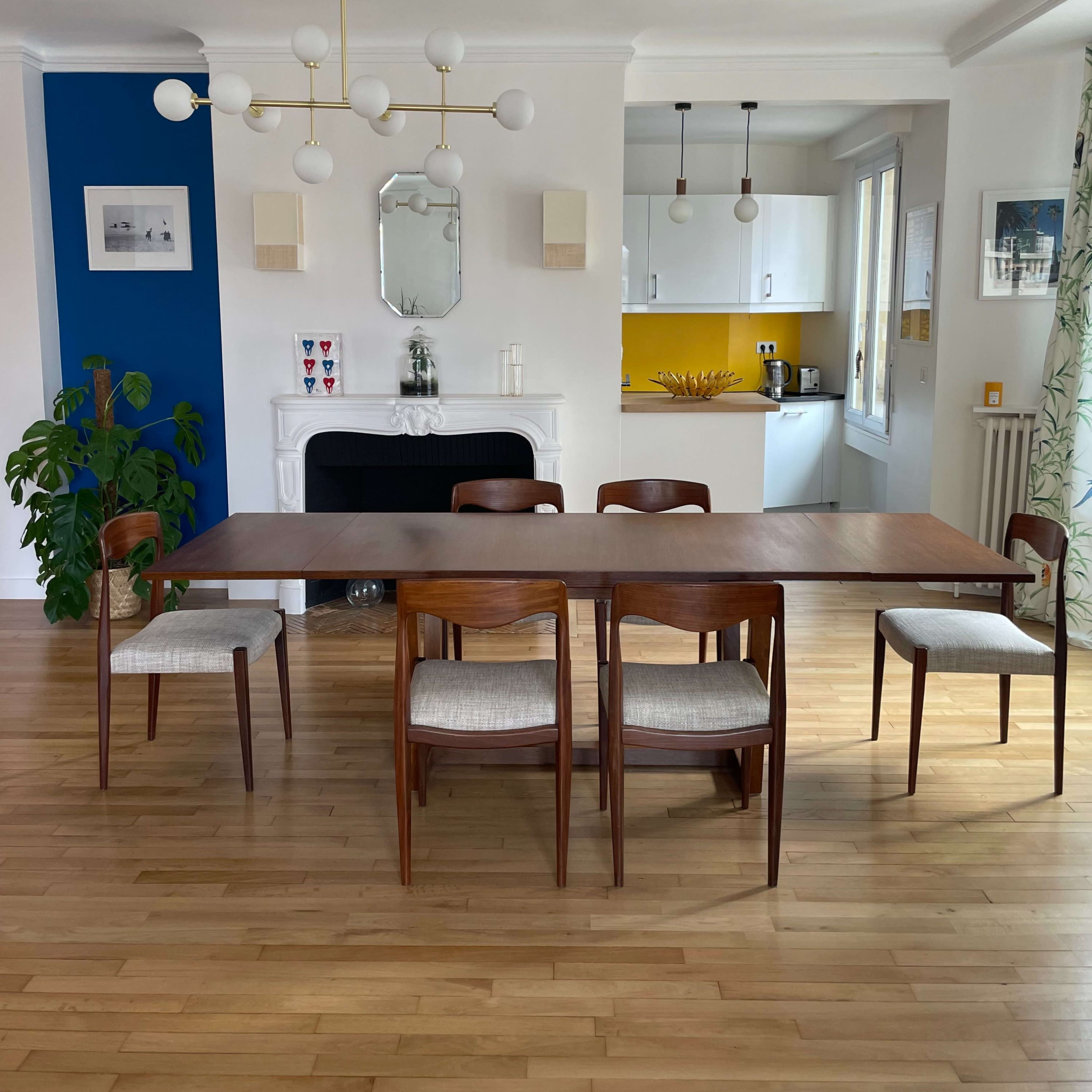 A wooden dining table is surrounded by four chairs in a bright room featuring a blue accent wall and a kitchen area visible in the background.