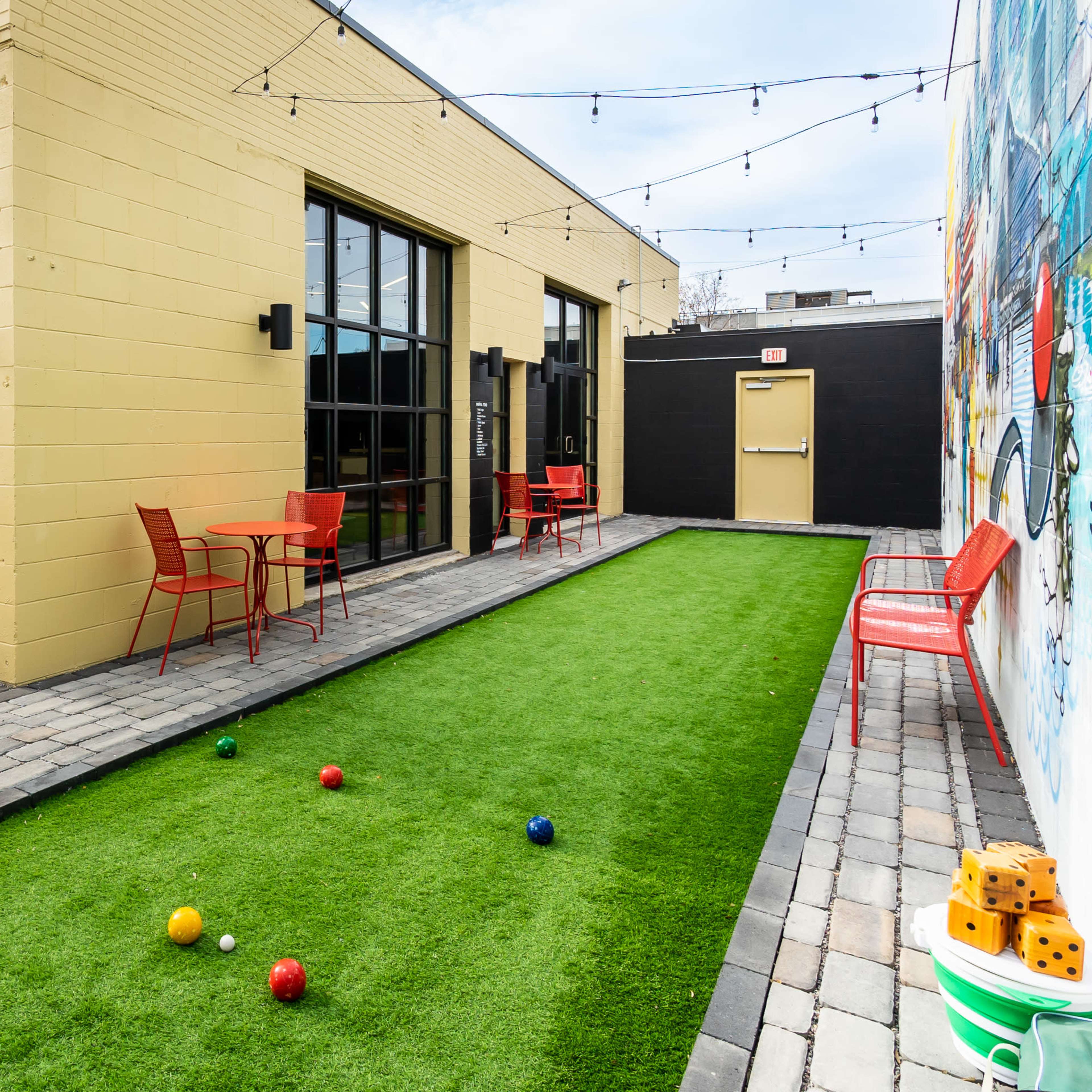 A courtyard features a green lawn with bocce balls, red chairs and tables, and a TV mounted on a yellow wall.