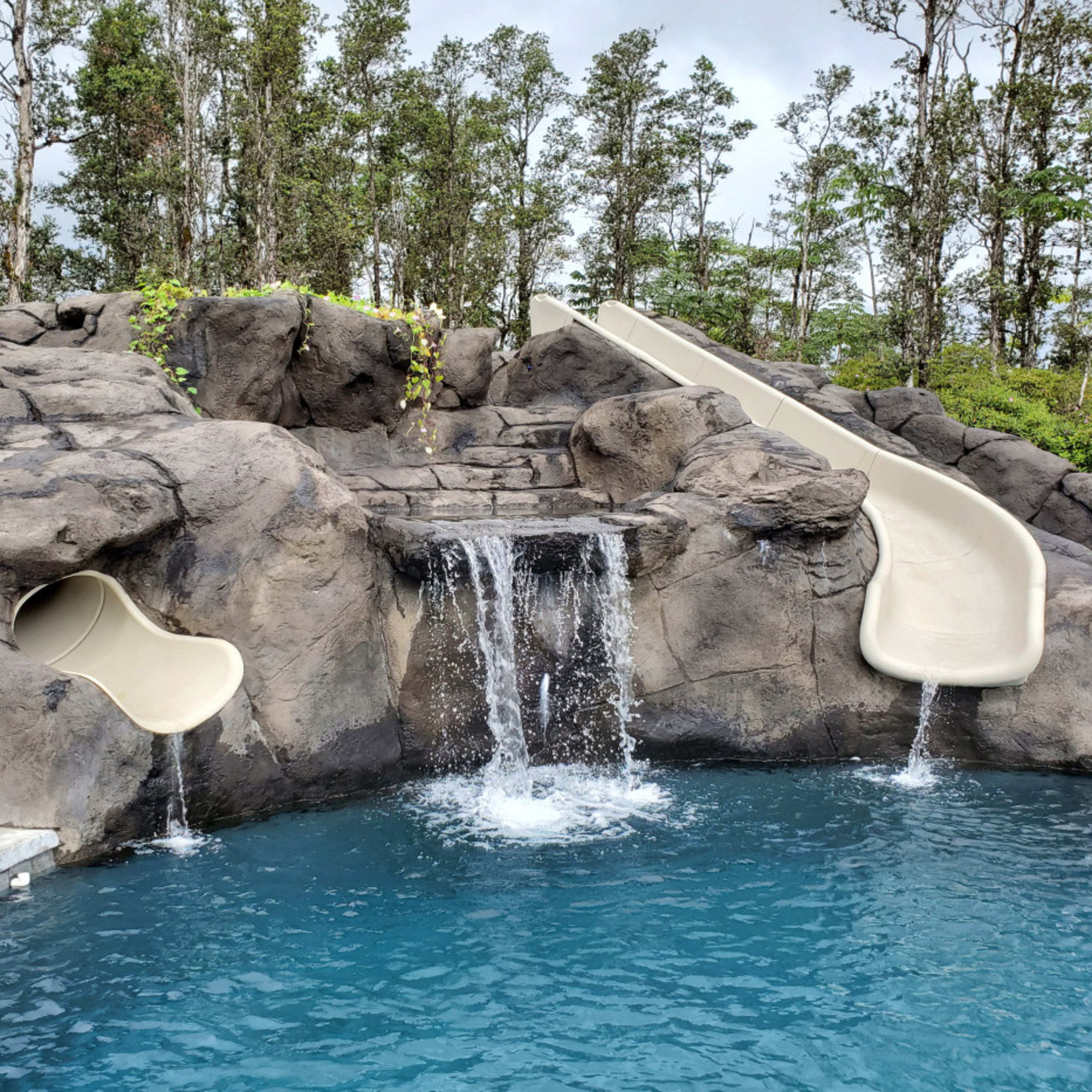 A rocky water slide structure next to a swimming pool, with two slides and a waterfall flowing into the pool area.