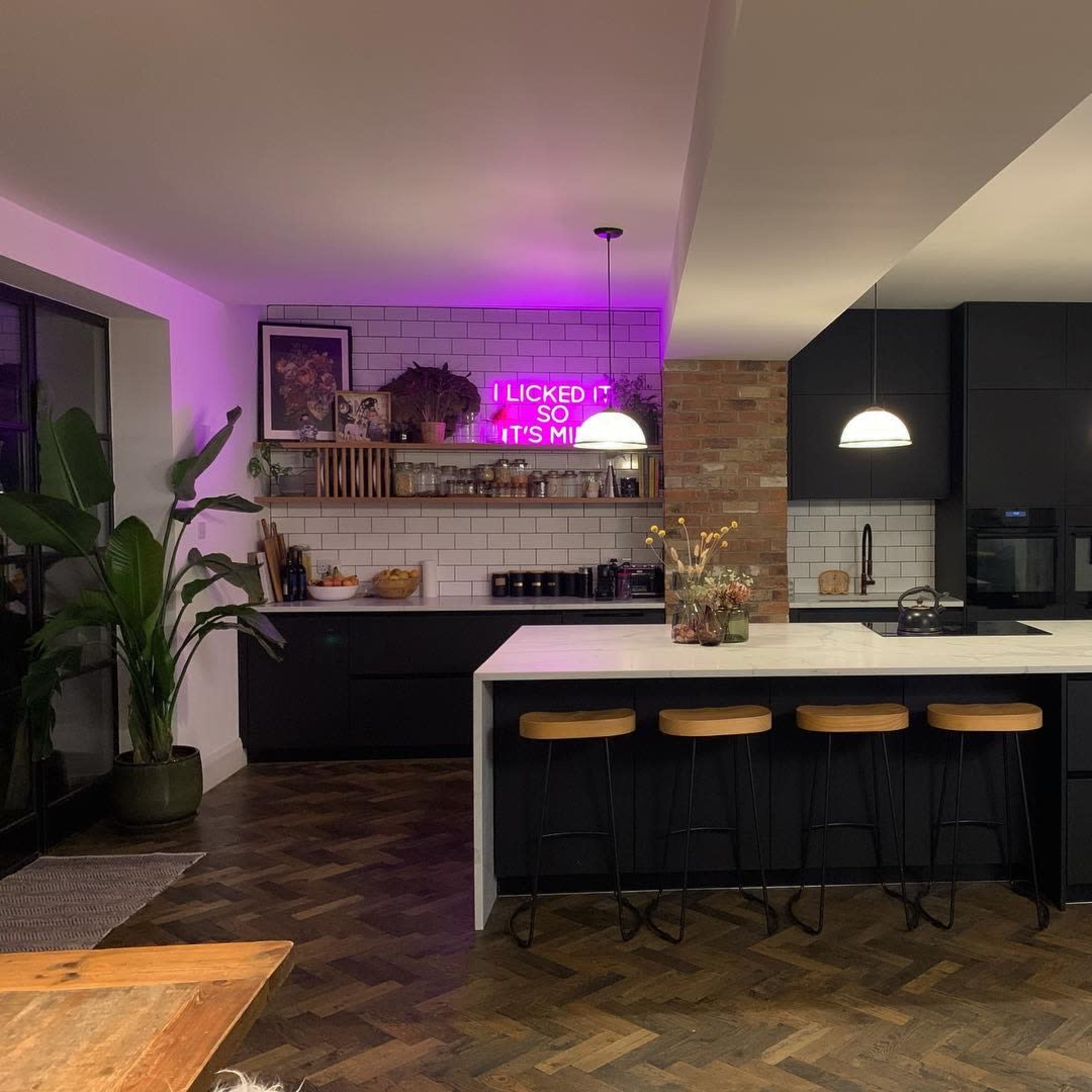A modern kitchen features a large marble island with black stools, dark cabinetry, and a decorative wall with plants and a neon light.