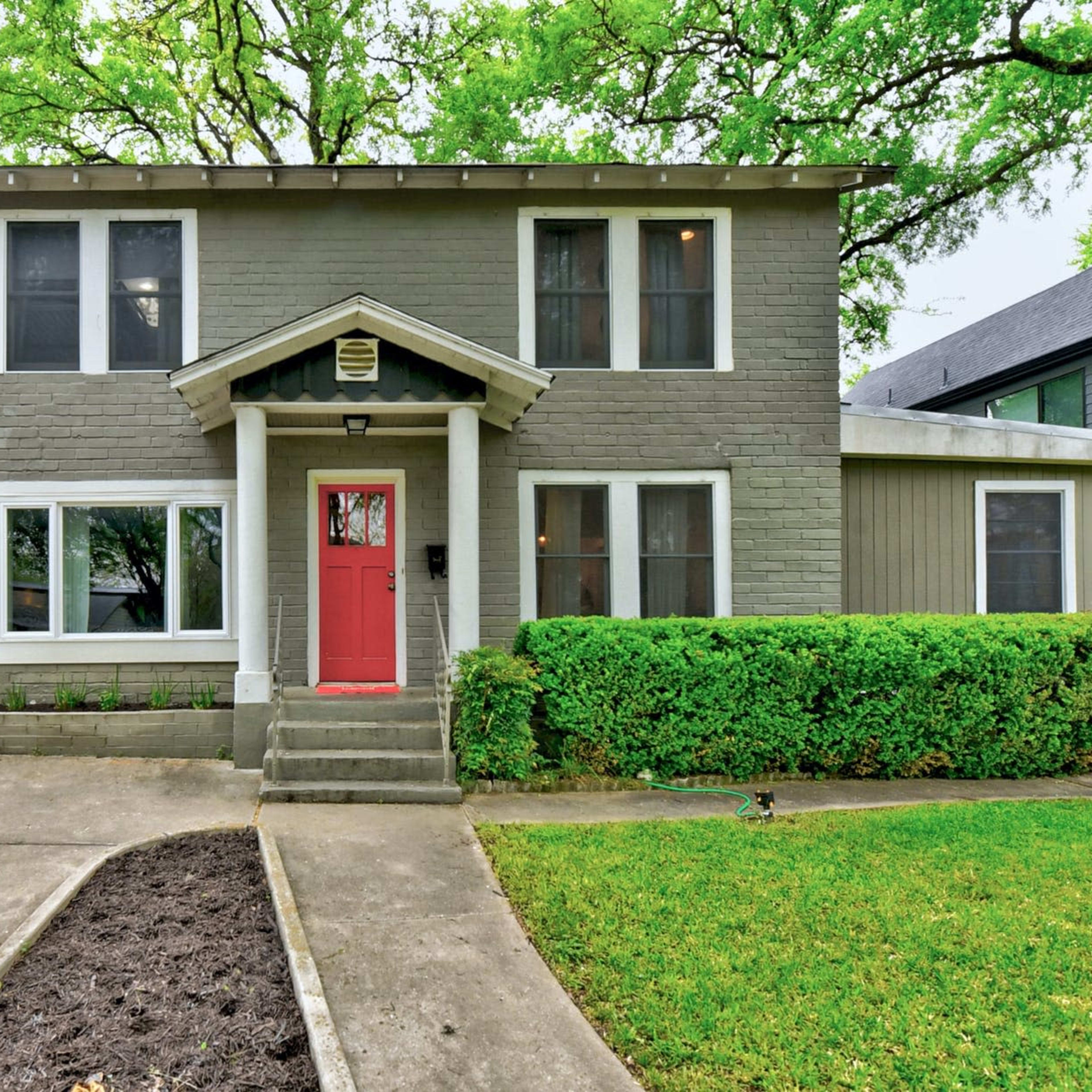 A two-story gray house with a red front door is surrounded by green foliage and has a carport on the left side.