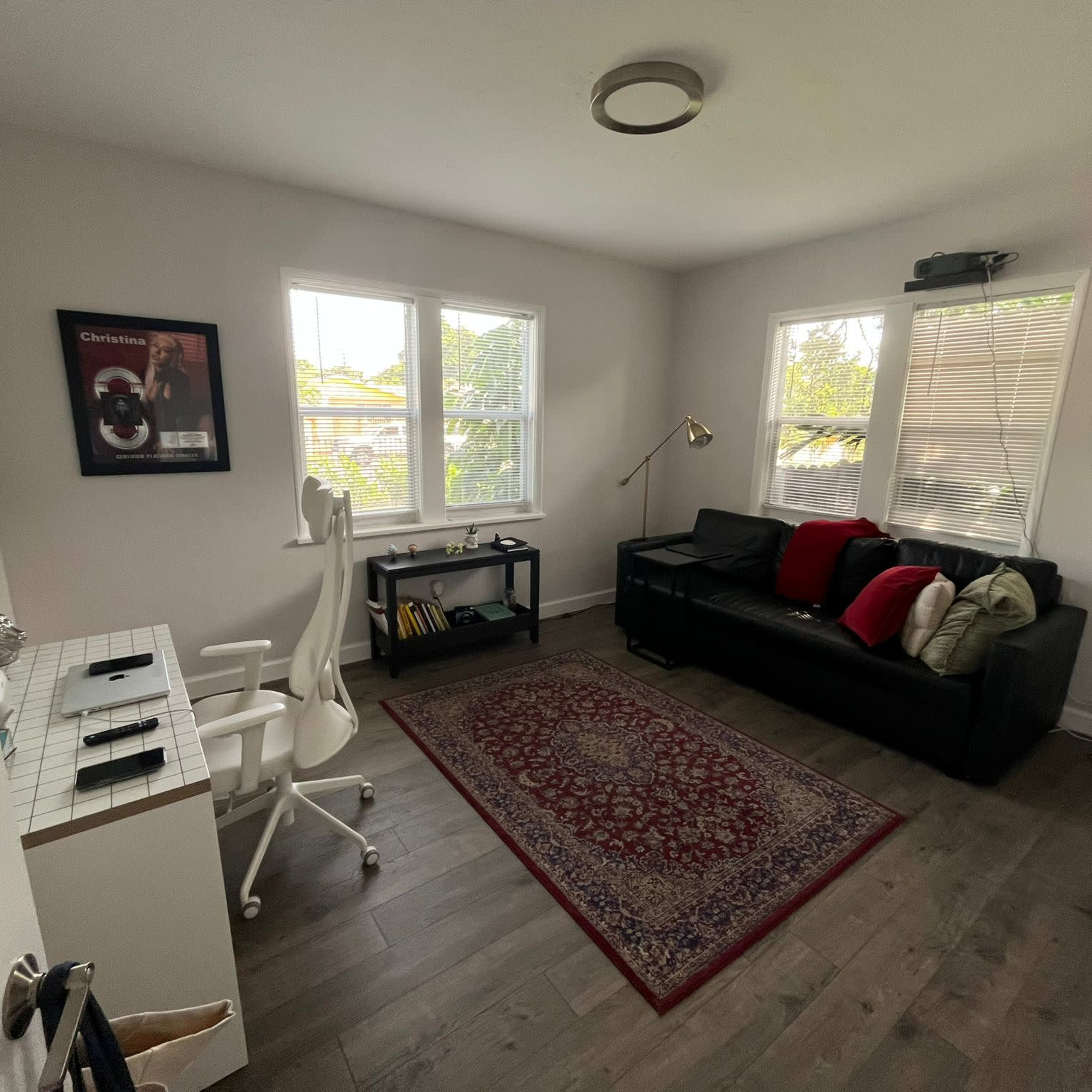 A well-lit office with a desk, ergonomic chair, and a black couch adorned with red pillows, featuring a patterned rug and two windows.