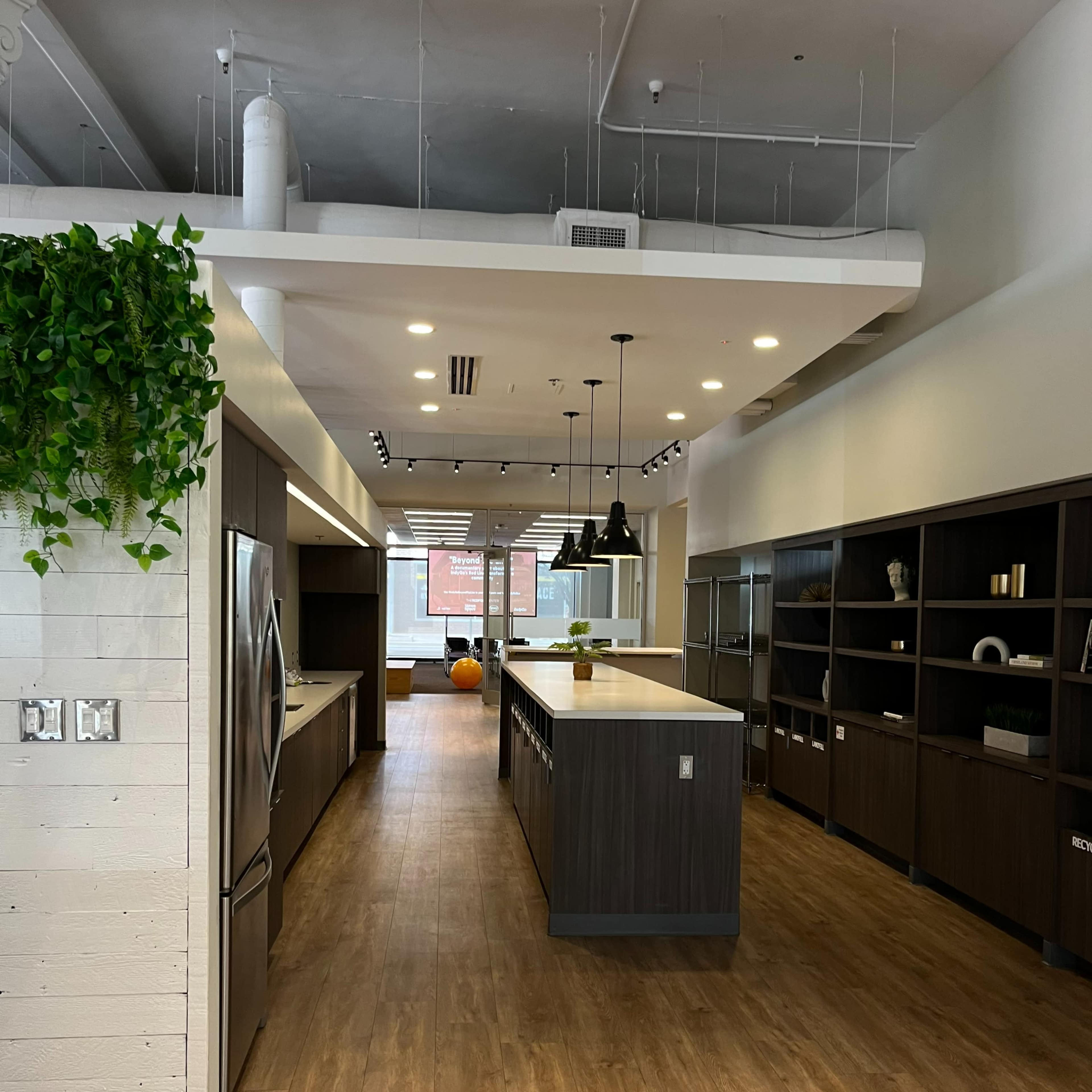 A modern kitchen with a long central island, dark cabinetry, overhead lighting, and large windows at the far end.