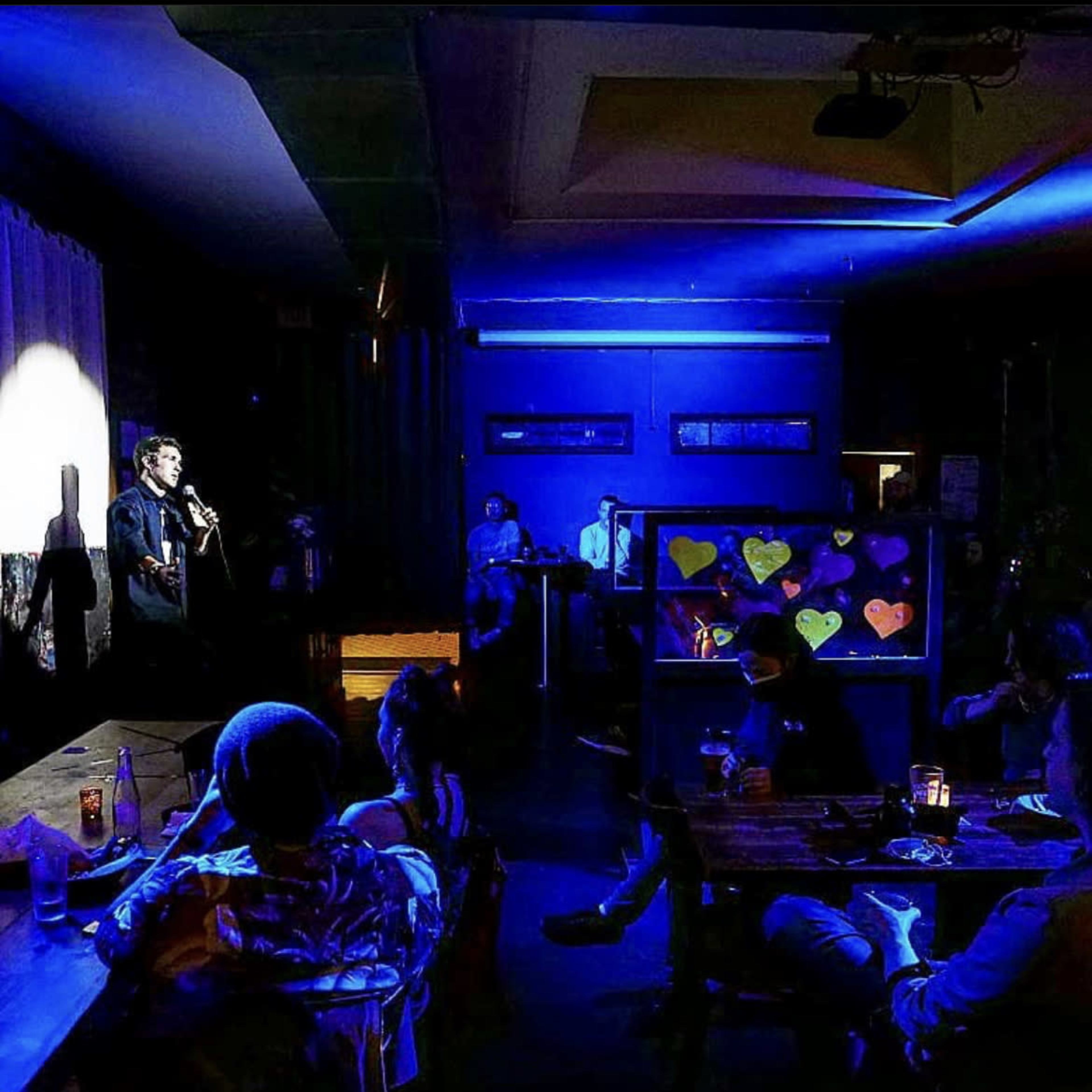 A comedian performs on stage in a dimly lit bar while an audience sits at tables, engaged in the show.