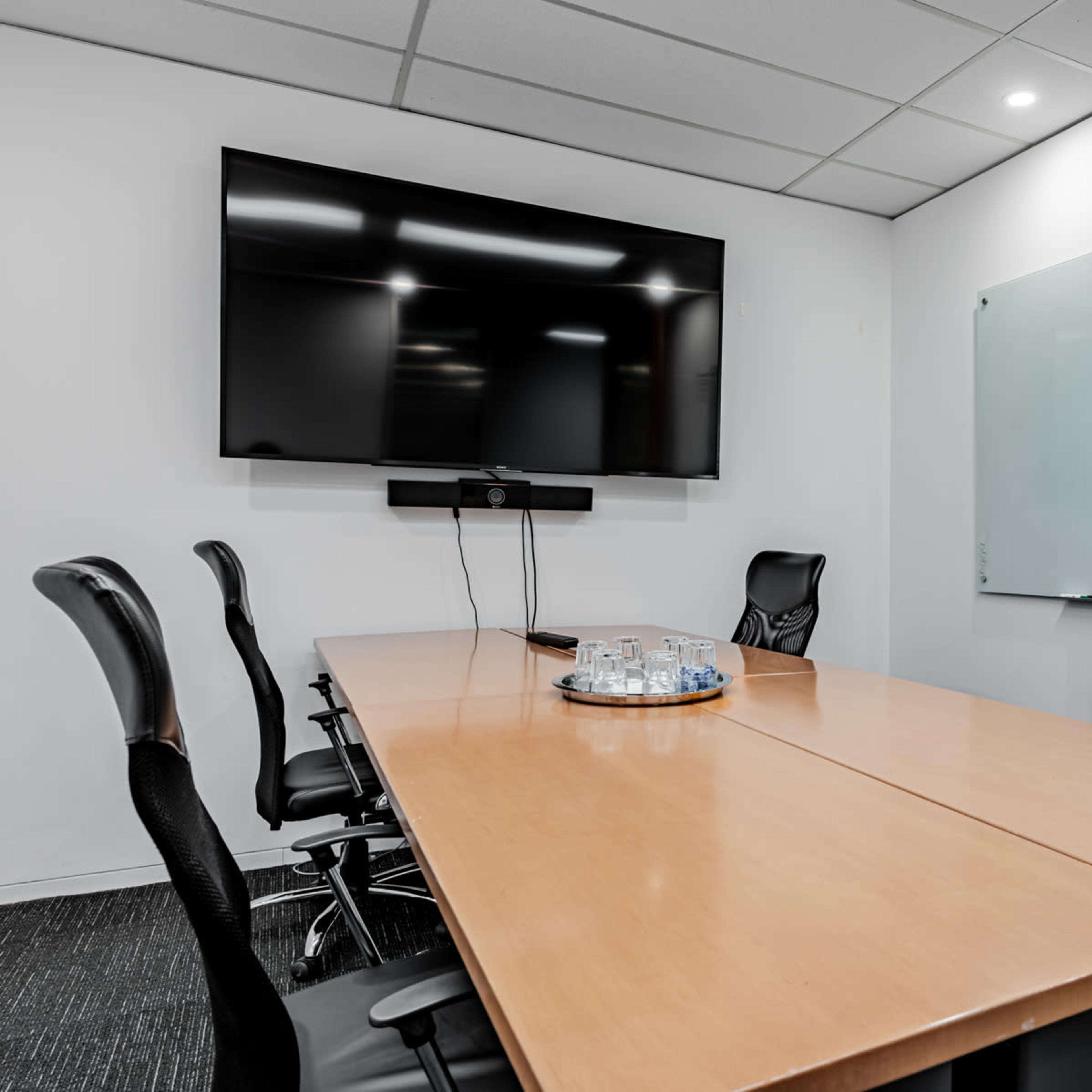 A modern conference room with a wooden table, six black leather chairs, a large wall-mounted television, and a whiteboard.