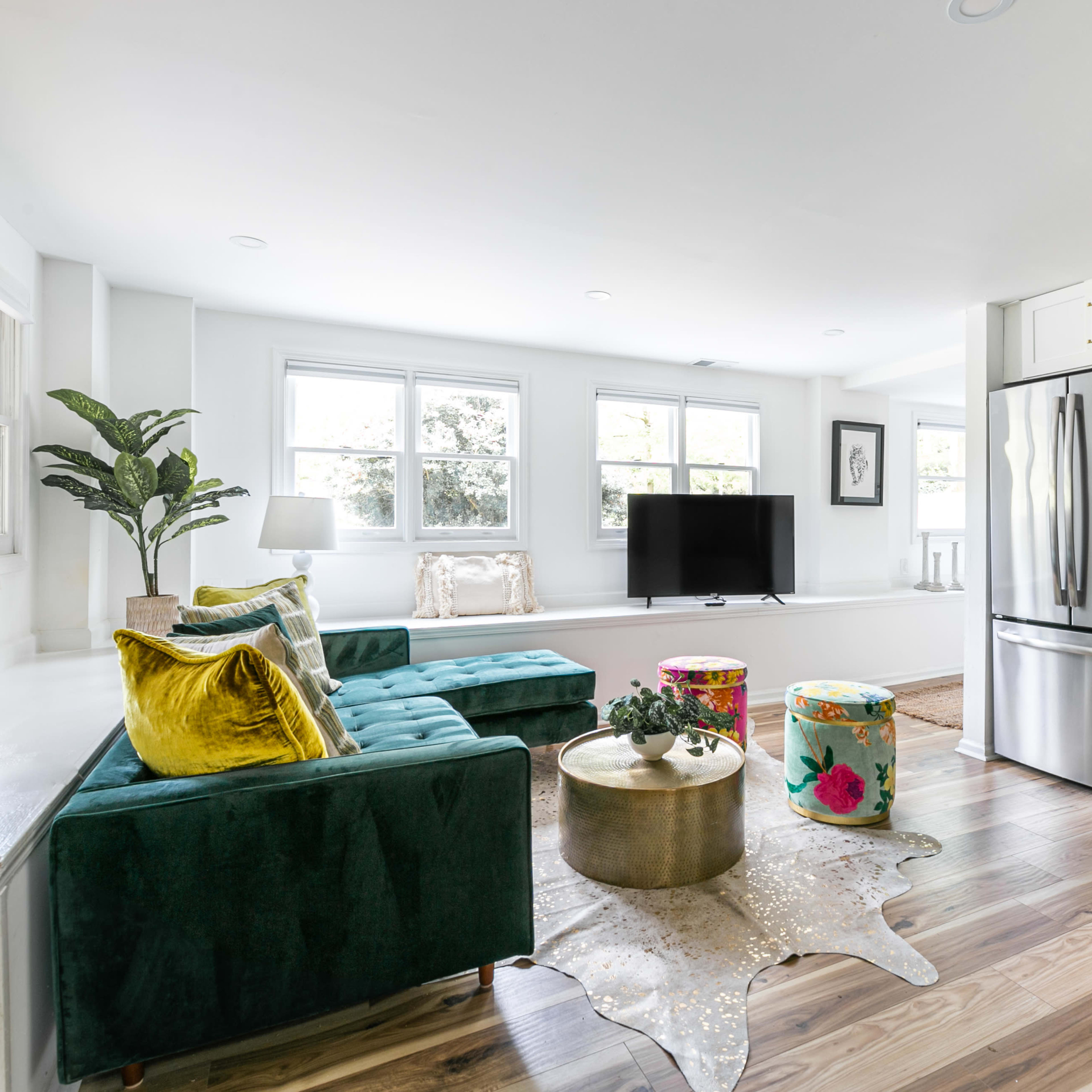 A modern living space features a green sectional sofa, a gold coffee table, colorful stools, and a kitchen area with stainless steel appliances.