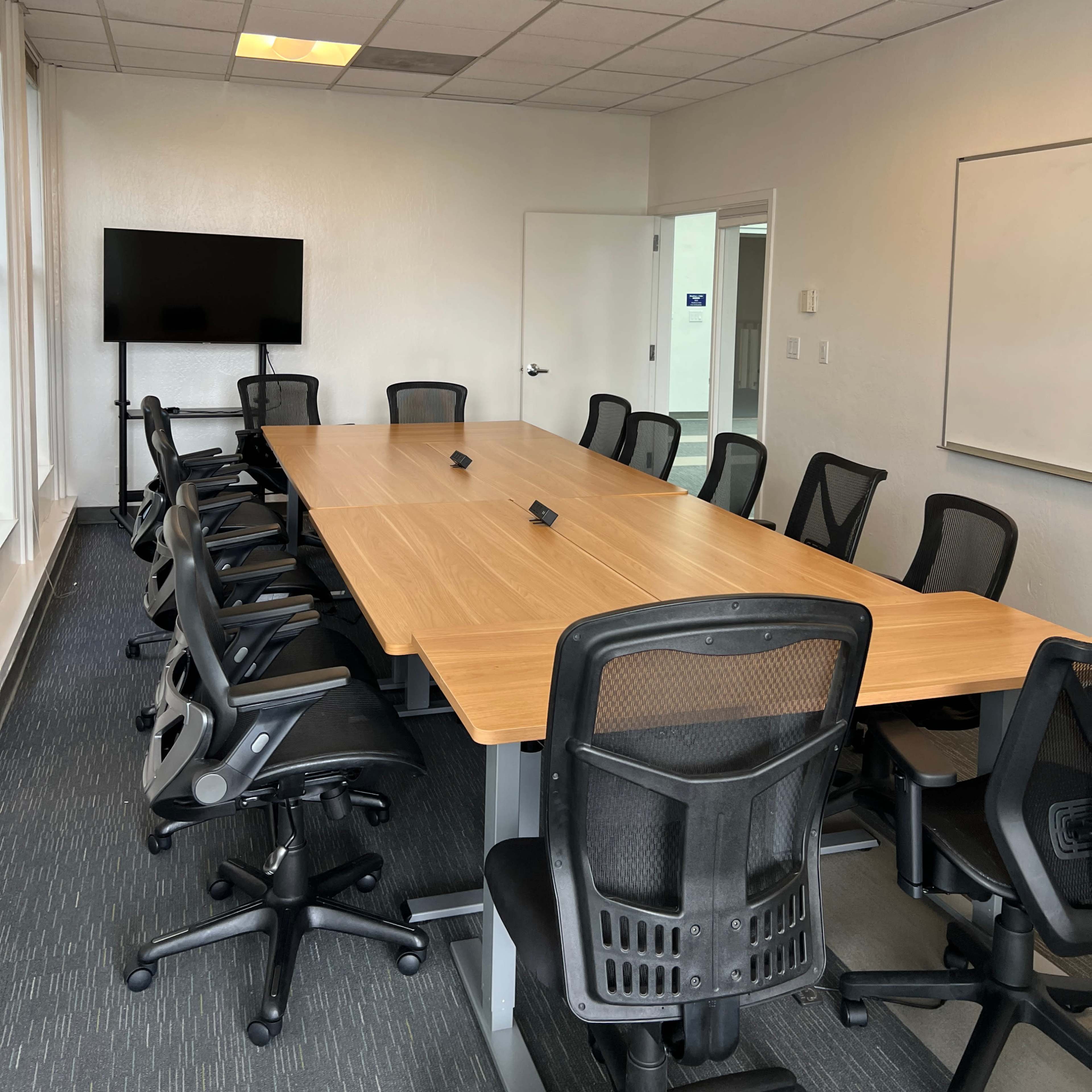 A spacious conference room furnished with a long wooden table surrounded by black office chairs, a television mounted on the wall, and a whiteboard visible in the background.