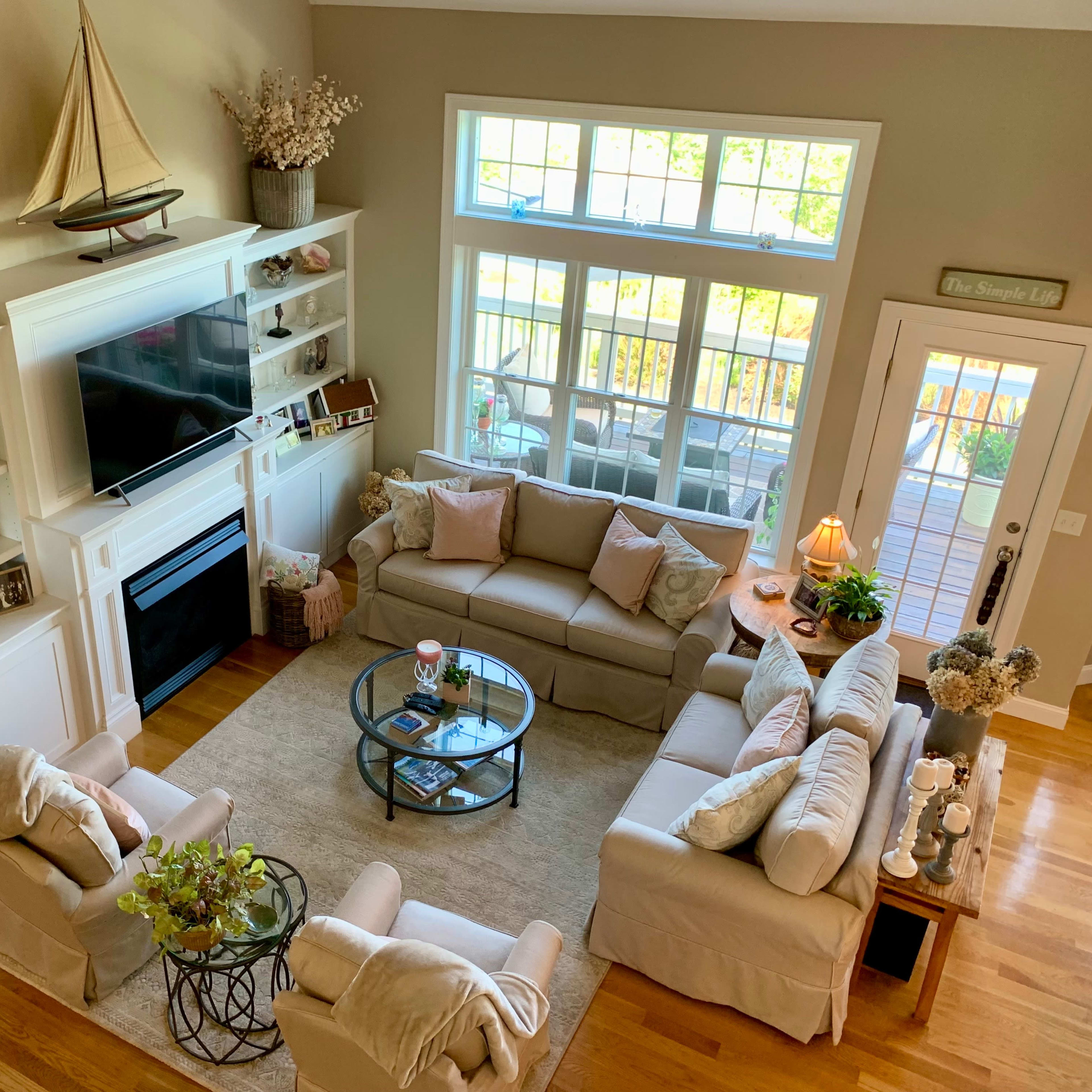 A well-lit living room features a central seating arrangement with a glass coffee table, surrounded by light-colored sofas and large windows showcasing a view outside.