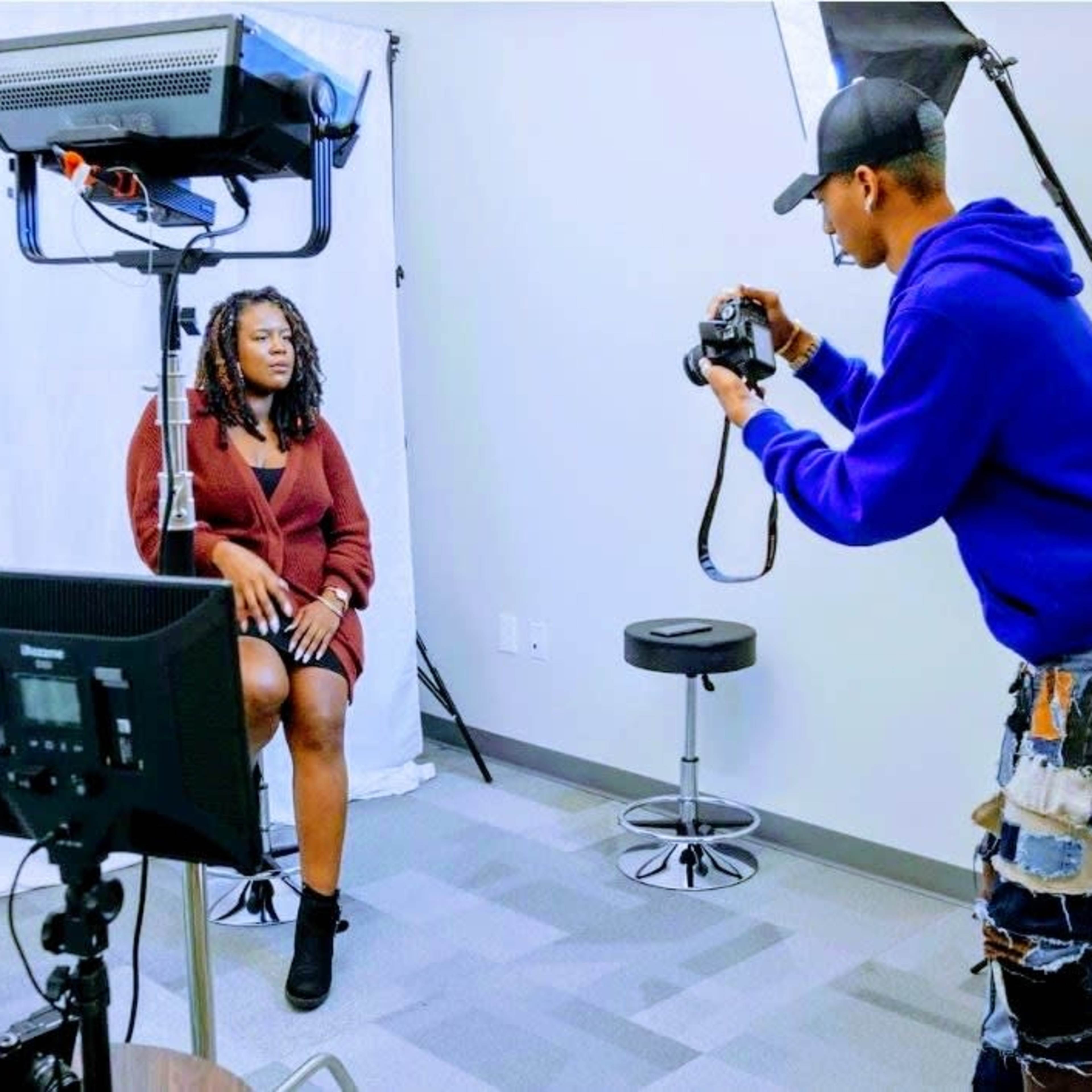 A photographer is taking a portrait of a woman seated in a studio equipped with professional lighting and a camera setup.