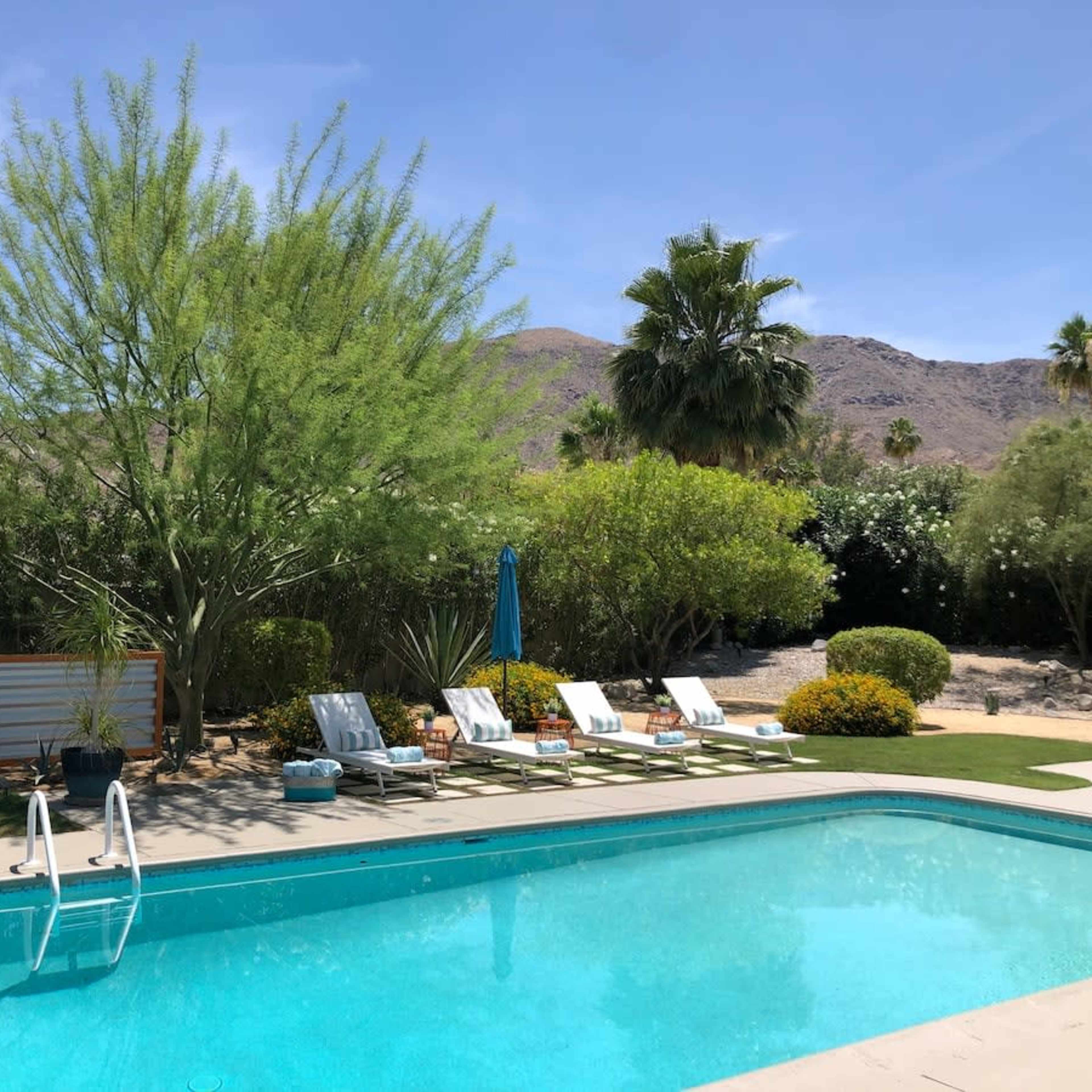 A swimming pool surrounded by lounge chairs, trees, and mountains in the background on a clear day.