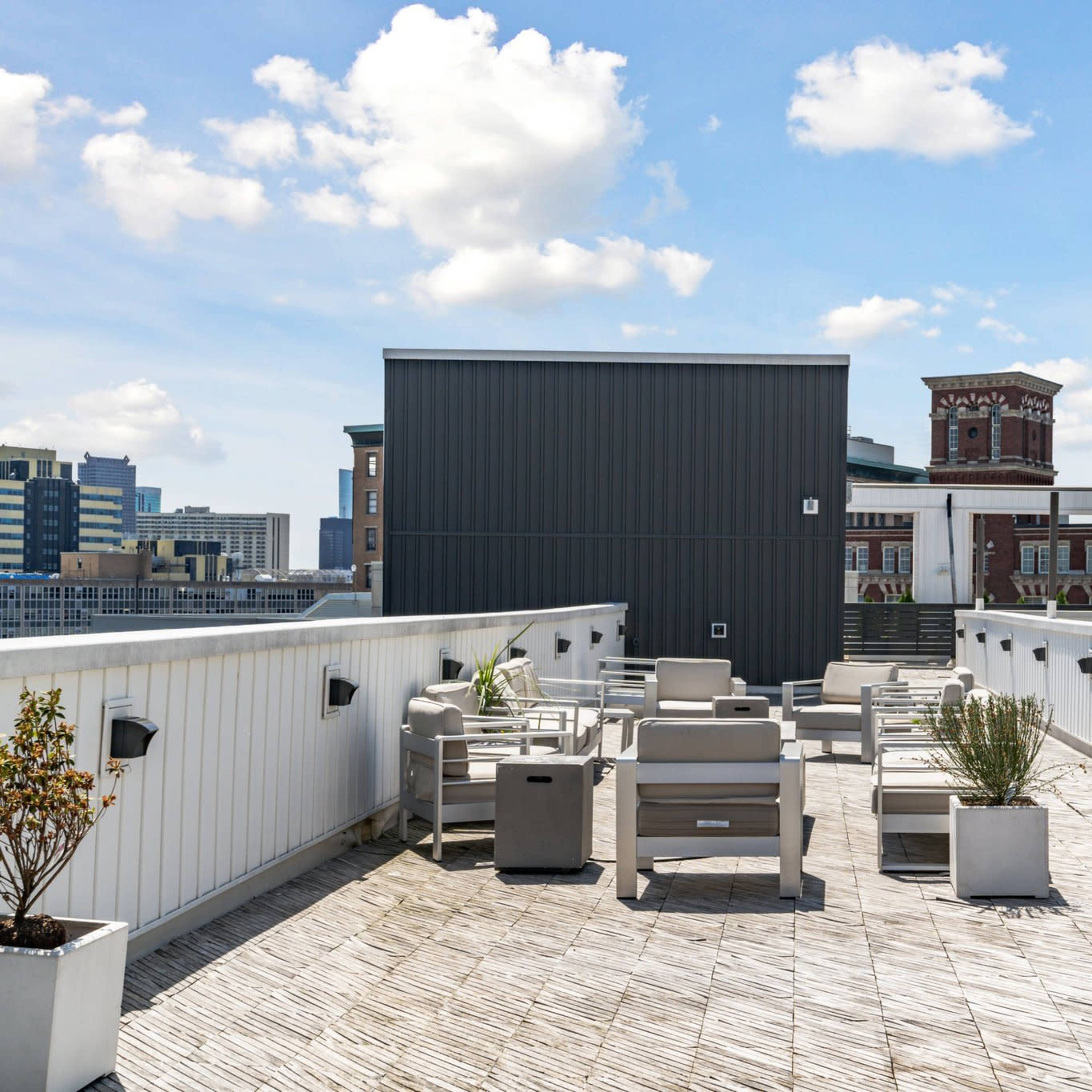 A rooftop terrace with seating areas and planters, overlooking a cityscape against a clear blue sky.