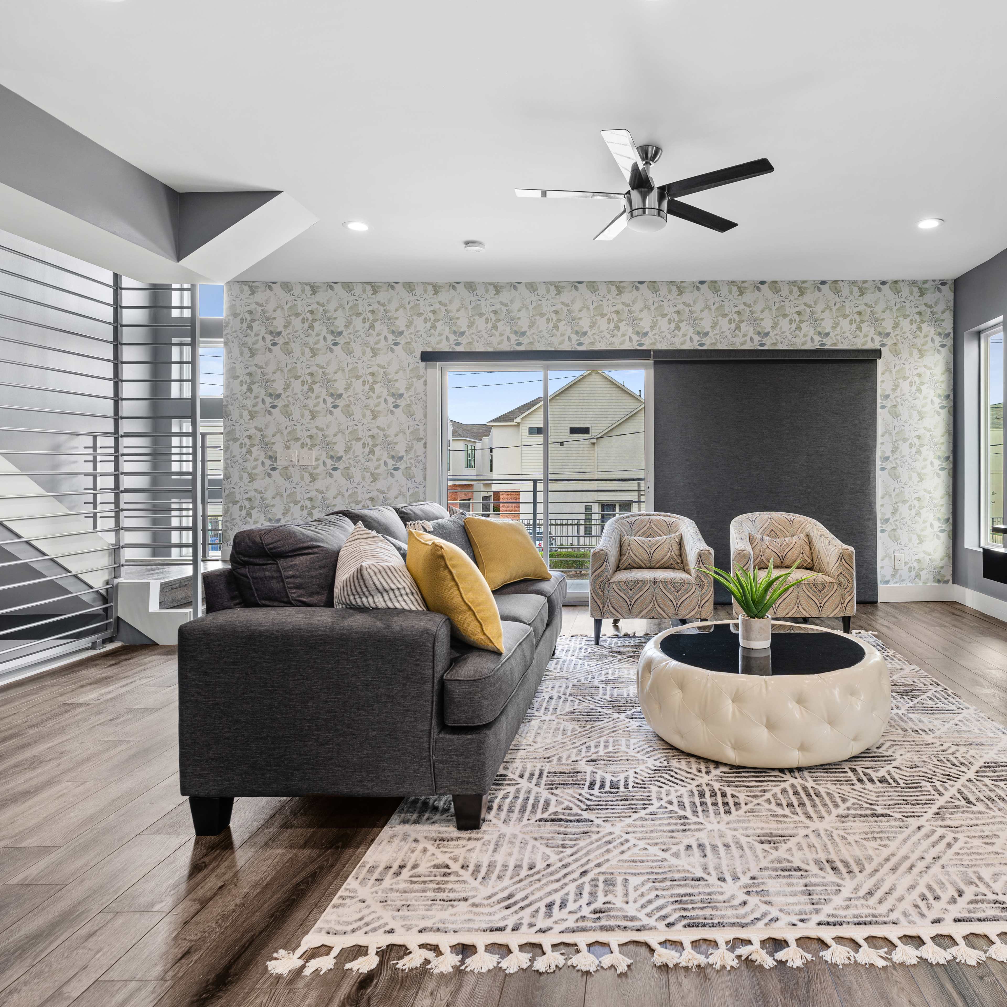 A modern living room features a gray sofa, two armchairs, a circular coffee table, and a large wall-mounted television, with decorative wallpaper and large windows.