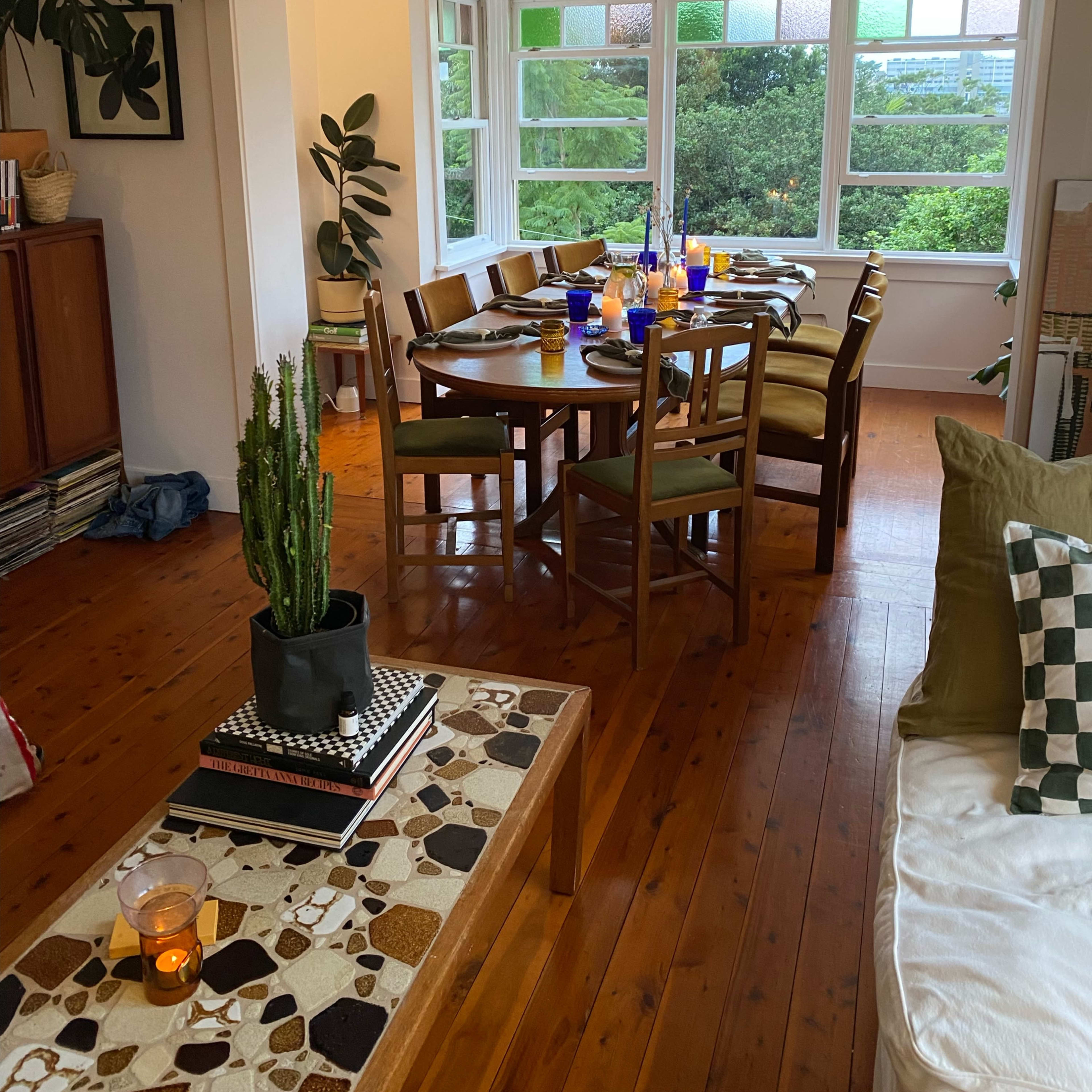A wooden dining table set for a meal is situated in a well-lit room with large windows and greenery visible outside.