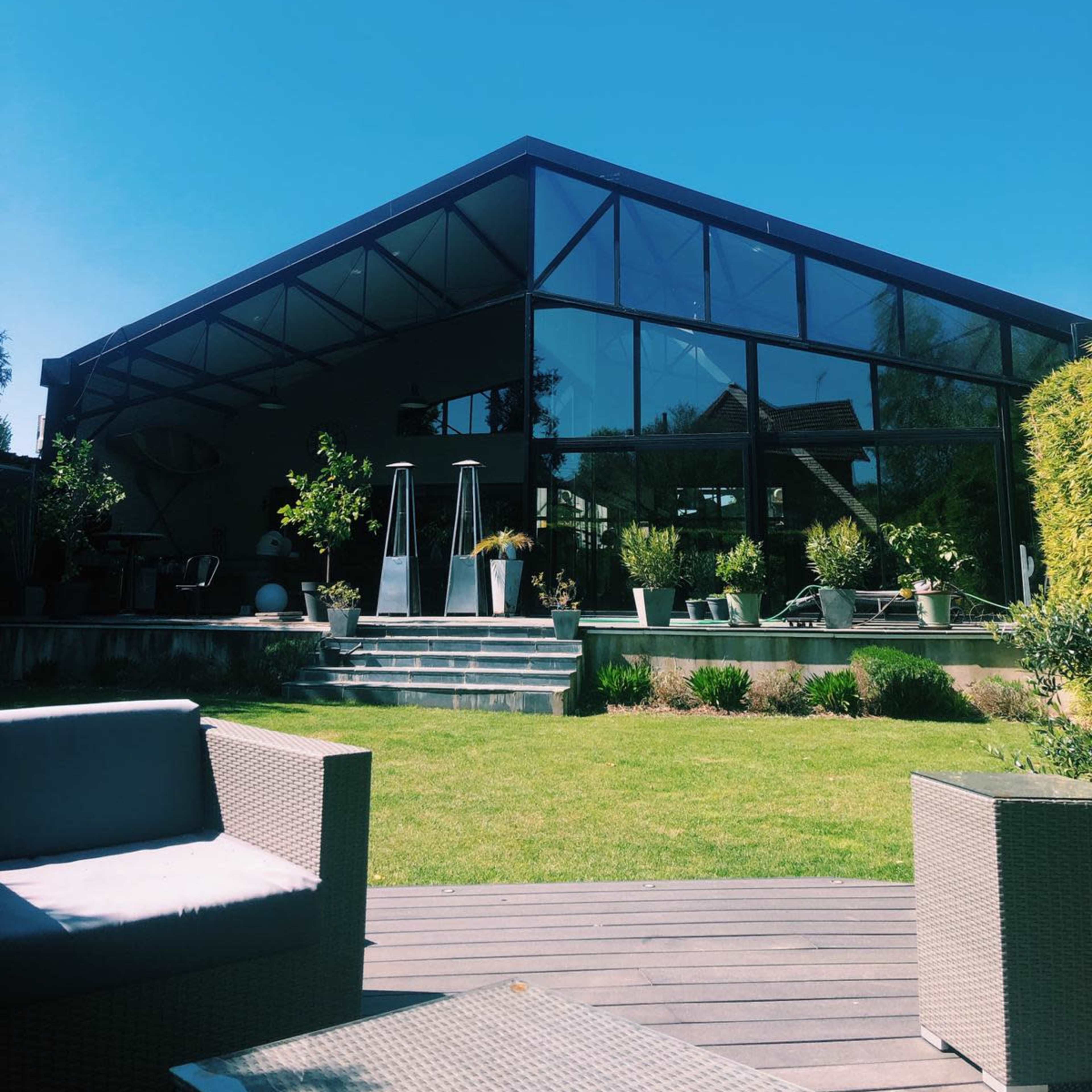 A modern glass-panel house with a sloped roof is surrounded by well-maintained greenery and outdoor seating.
