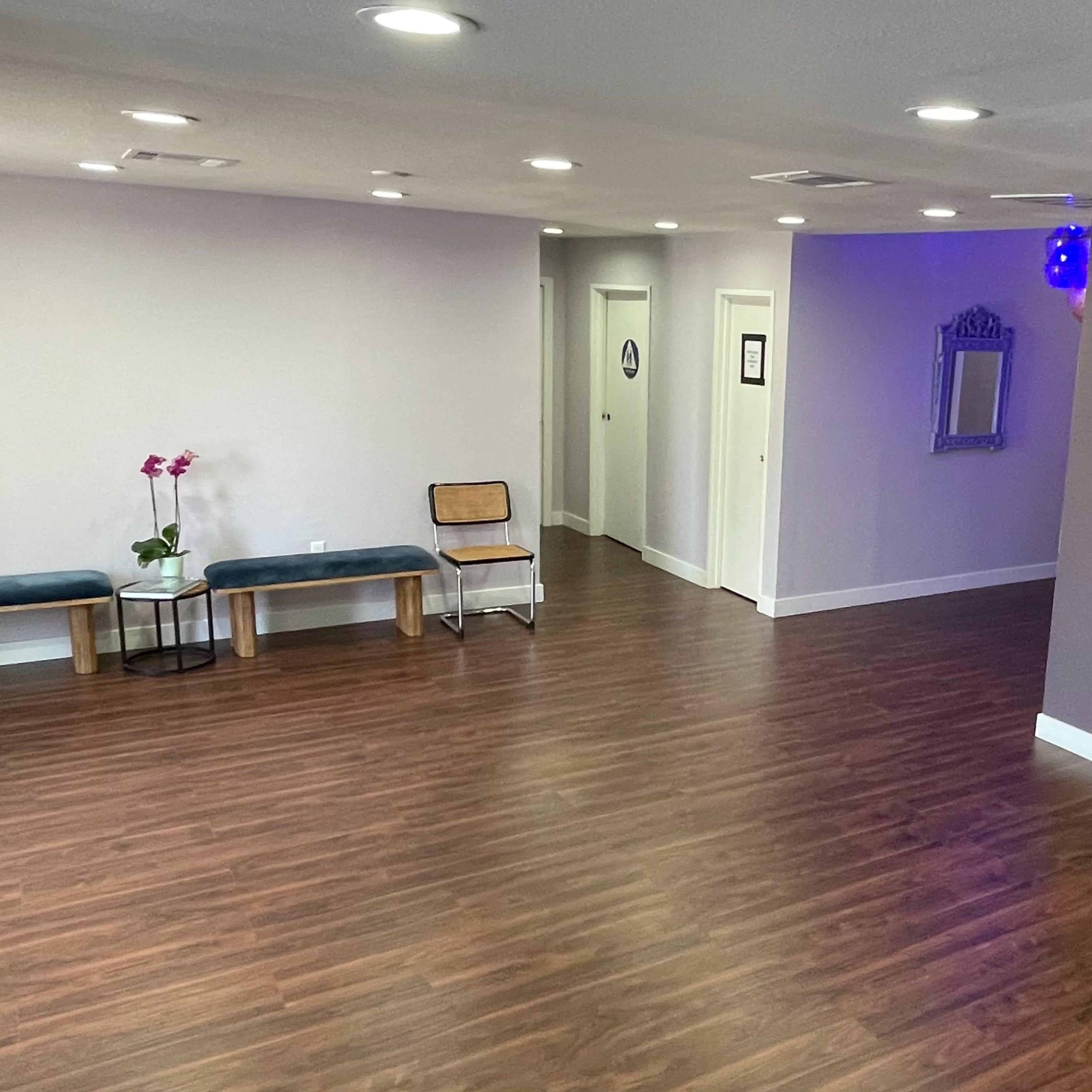 A spacious, well-lit interior with wooden flooring, featuring two benches, a television on the wall, and a hallway leading to closed doors.