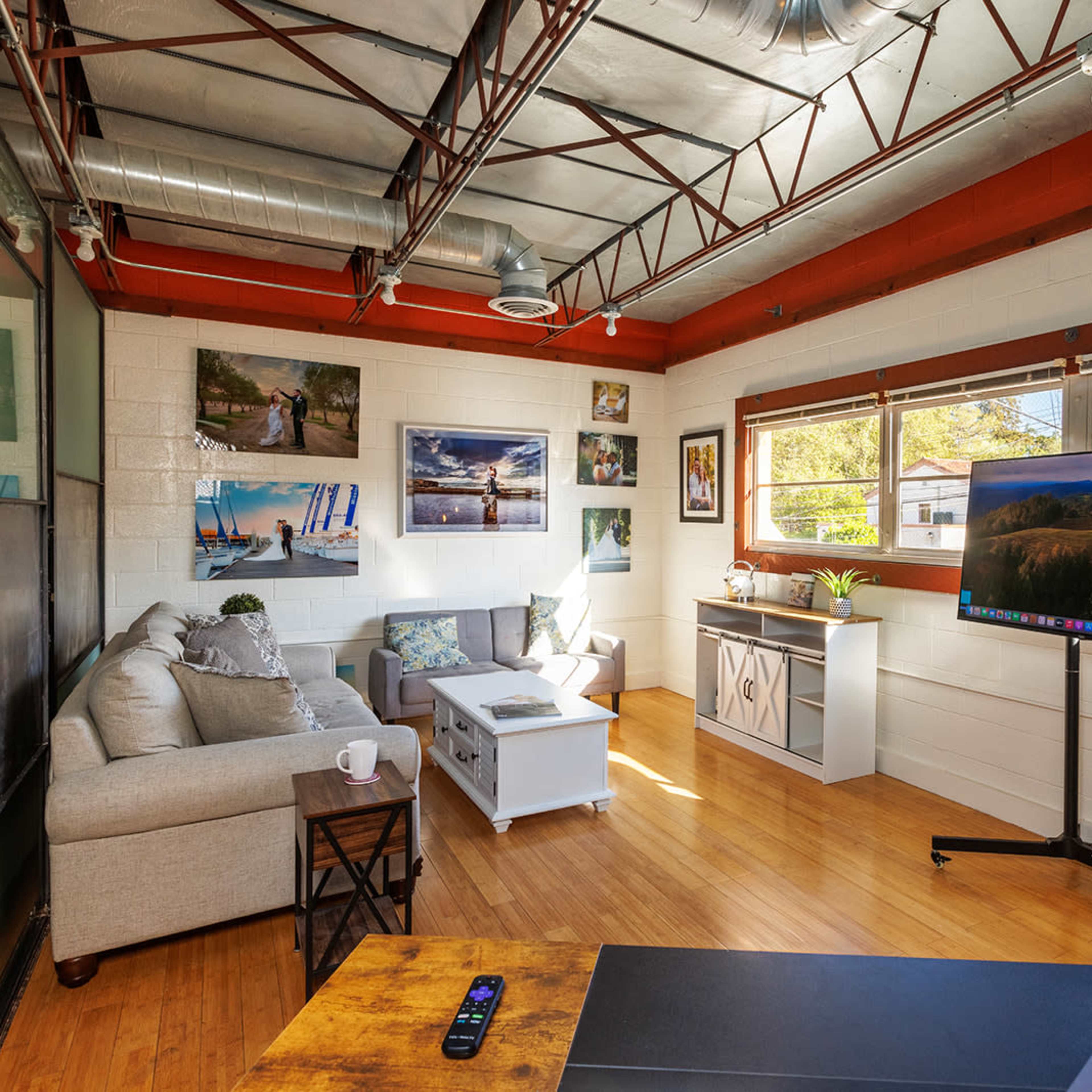 A modern office space featuring a sofa, a coffee table, a large monitor on a stand, and several framed photographs on the walls.