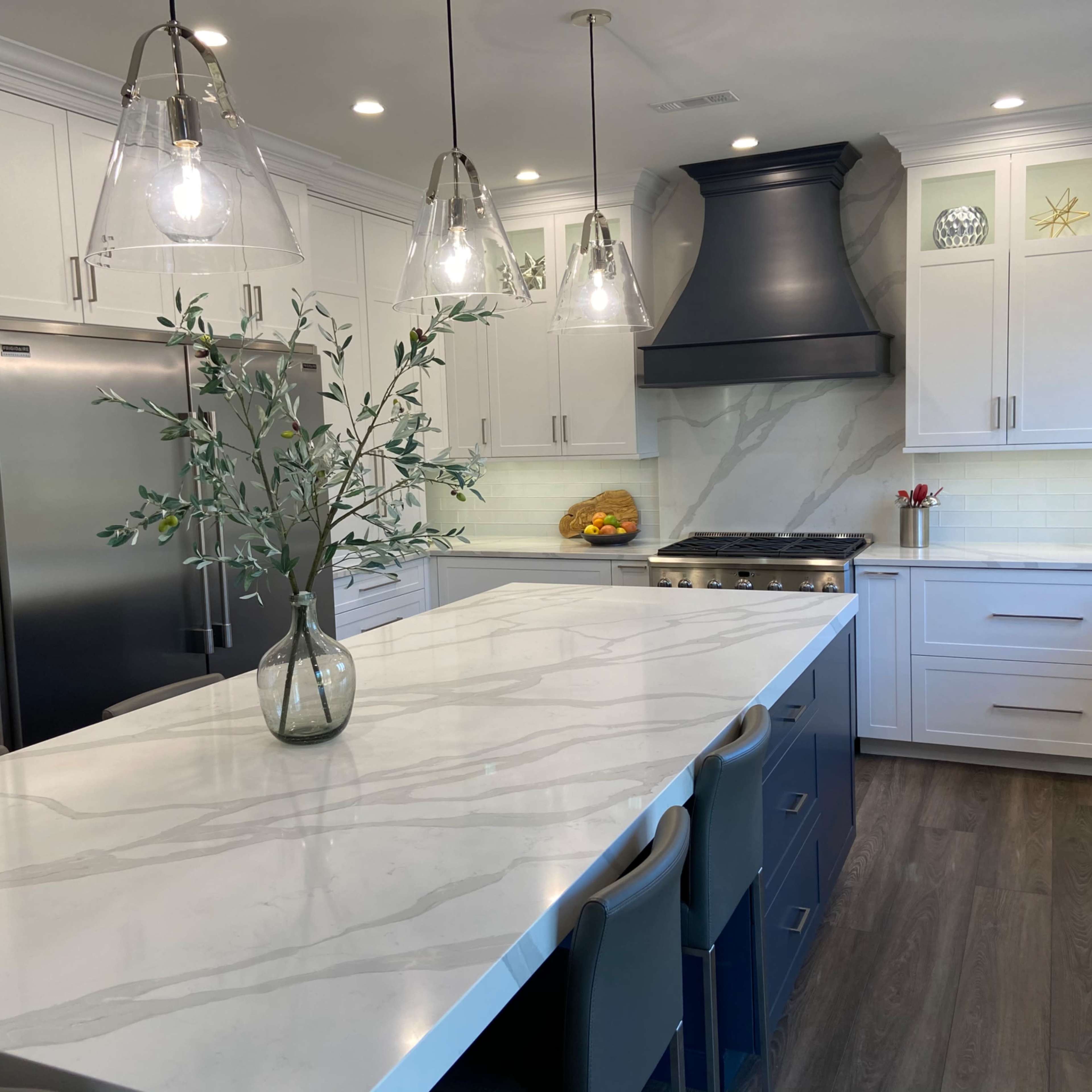 A modern kitchen features a large island with a marble countertop, white cabinetry, stainless steel appliances, and pendant lighting.