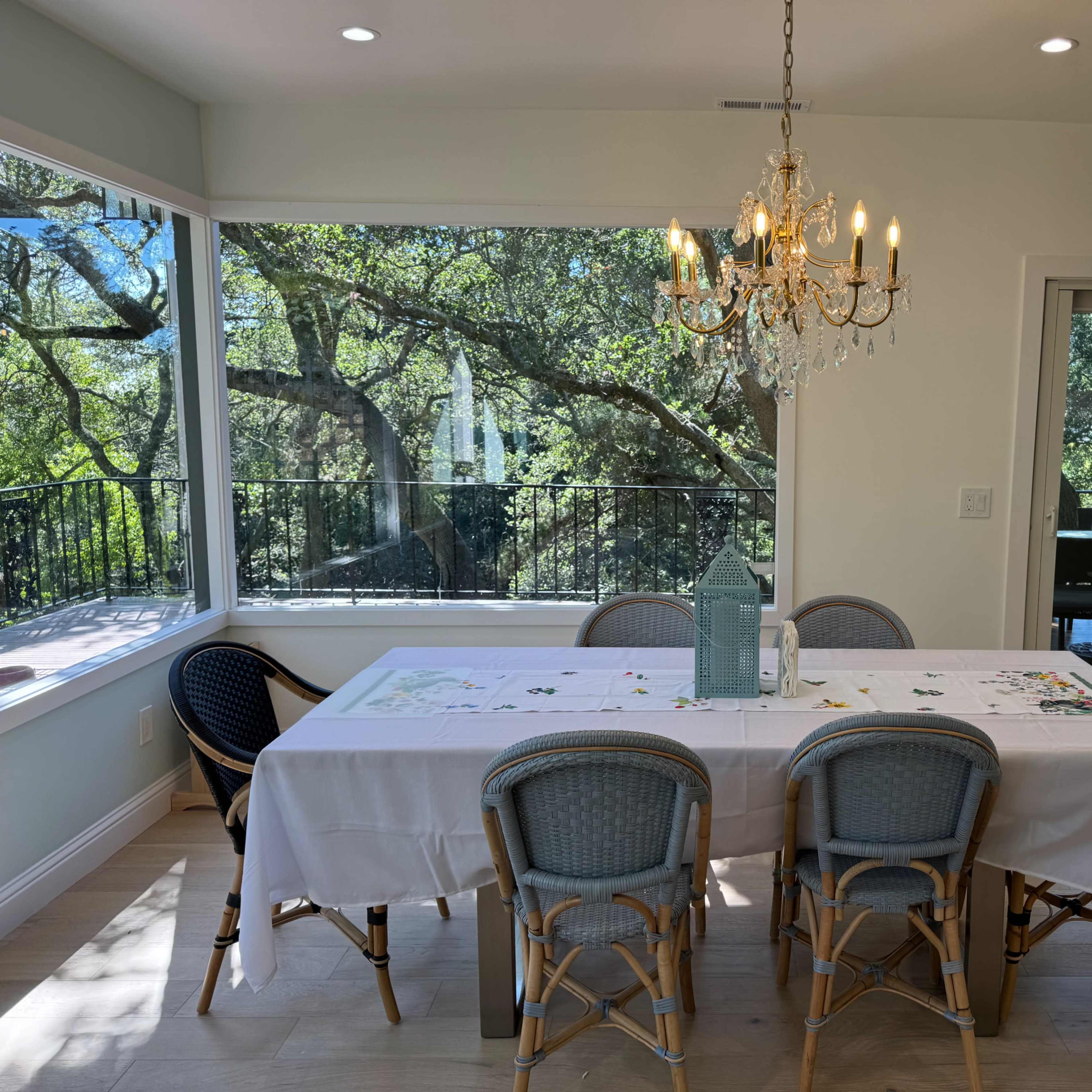 A dining area features a long white table surrounded by eight chairs, with a chandelier overhead and large windows showcasing a view of trees outside.